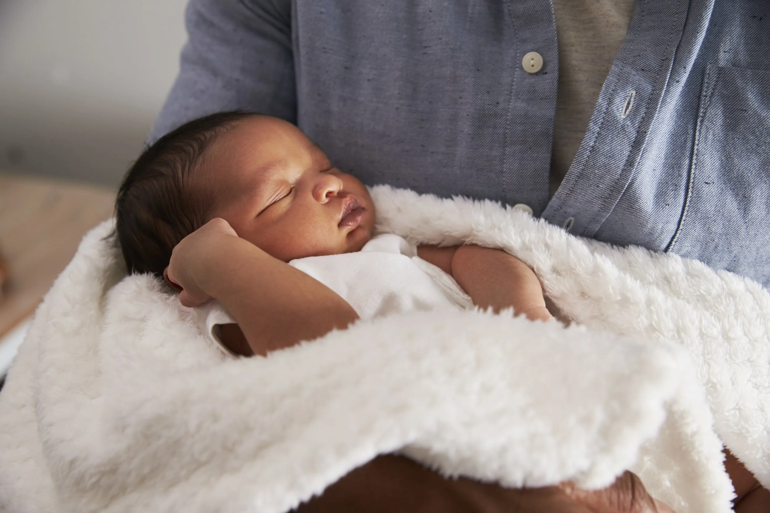 A sleeping newborn baby held gently by an adult, wrapped in a soft white blanket, with the adult wearing a grey shirt and a cap.
