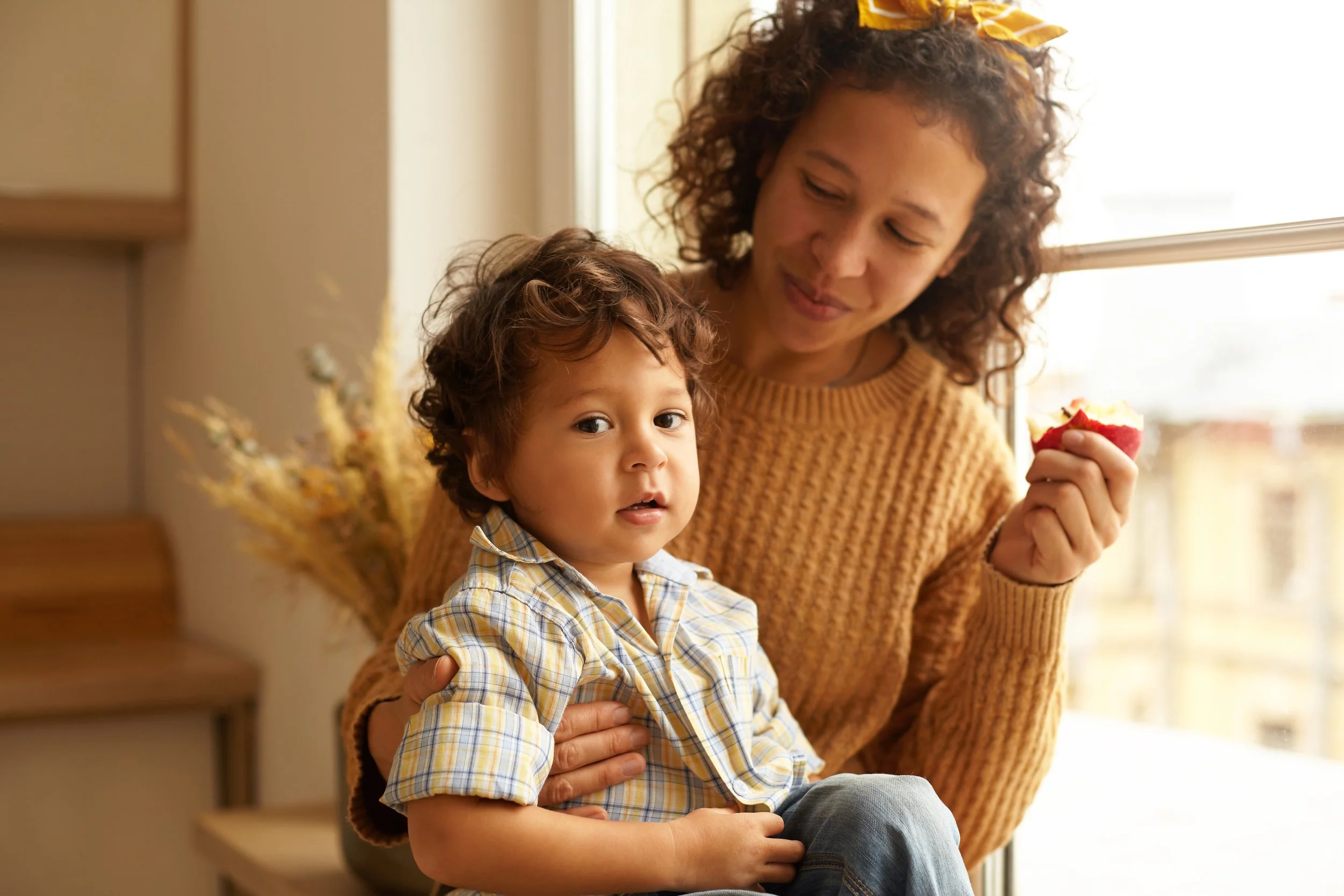 A woman with curly hair and a yellow headband holding a young boy with curly hair and a plaid shirt, sitting near a window with sunlight.