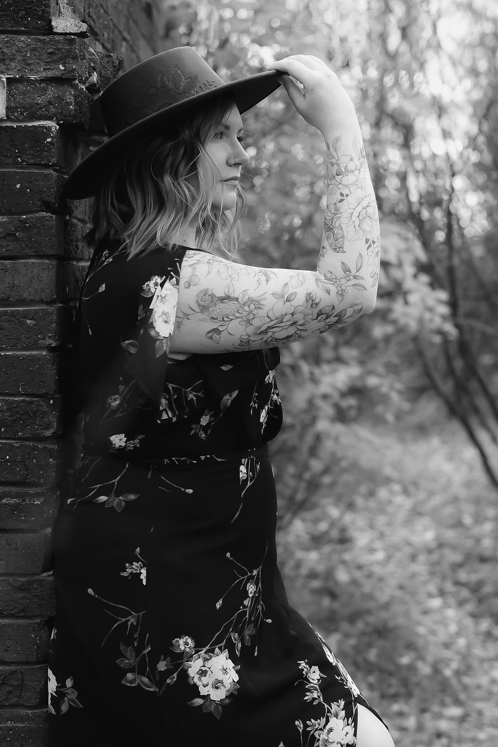 A woman with wavy hair, tattoos on her arm, wearing a floral dress and a wide-brimmed hat, stands beside a brick wall in a natural outdoor setting.