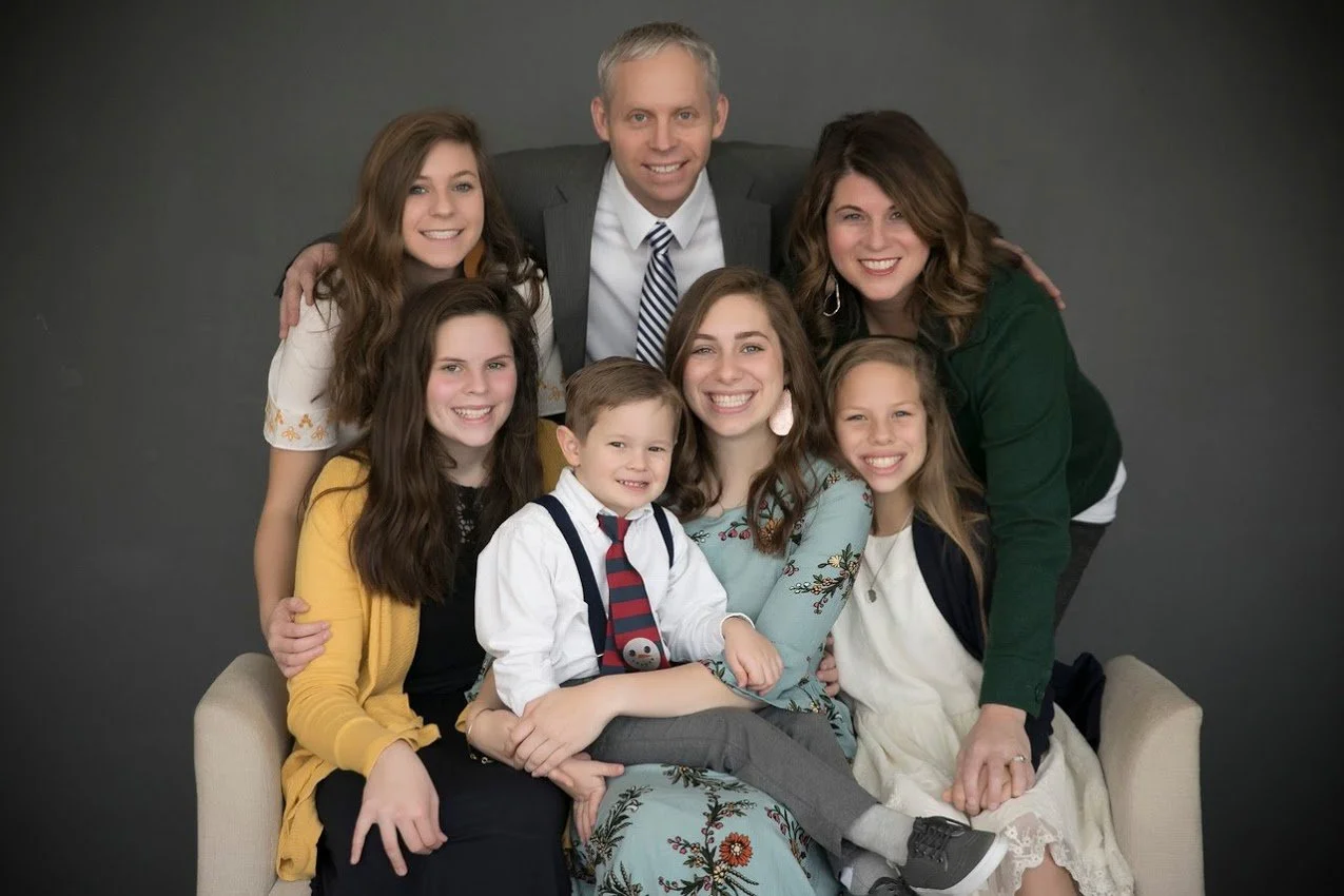 A multi-generational family portrait with six people, including three women, a man, and two children, smiling and sitting close together on a beige sofa against a plain gray background.