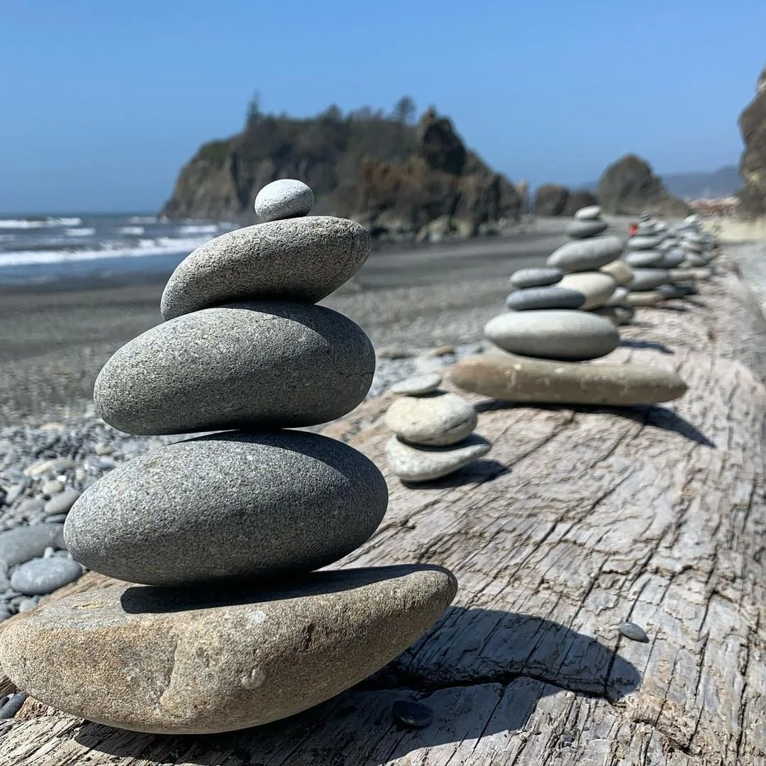 Line of stacked smooth stones on weathered driftwood at the beach with rocky cliffs and blue sky in the background.