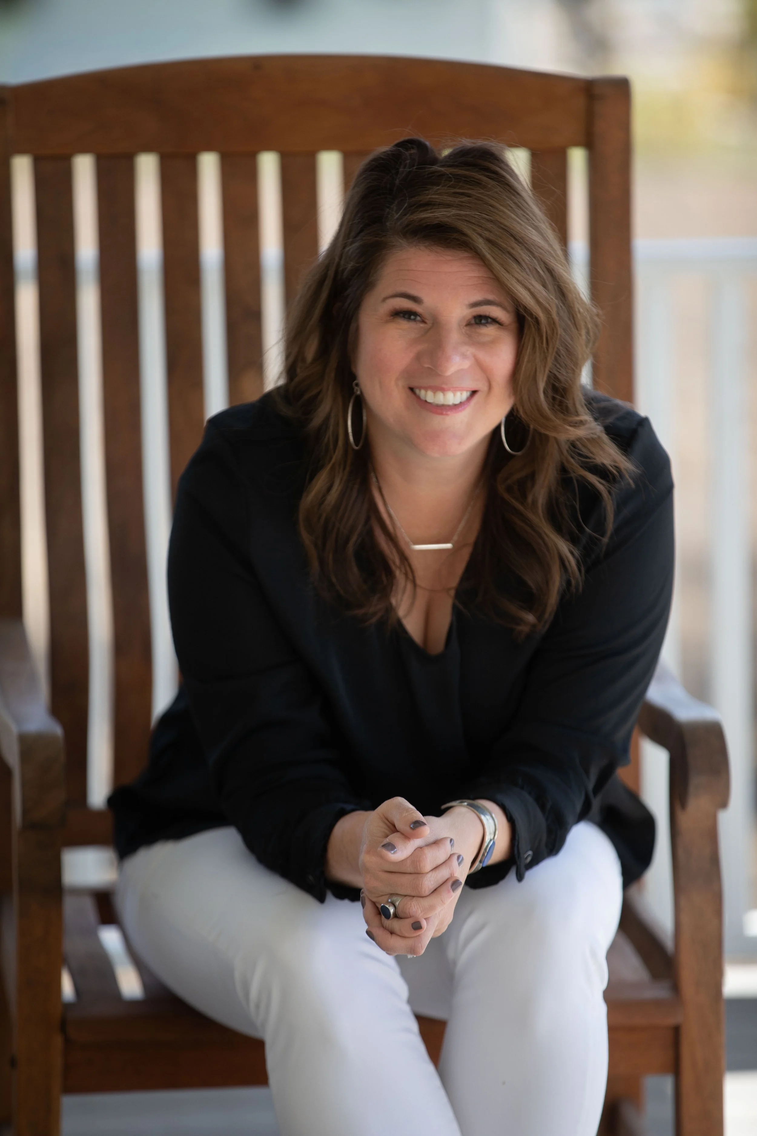 A woman with wavy brown hair sitting on a wooden chair, smiling with her hands clasped together, wearing a black top and white pants.