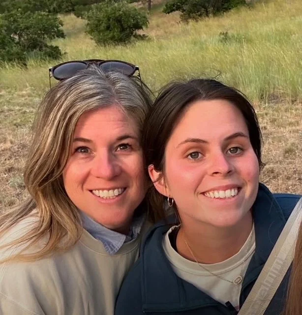 Two smiling women taking a selfie outdoors in a grassy area with trees in the background.