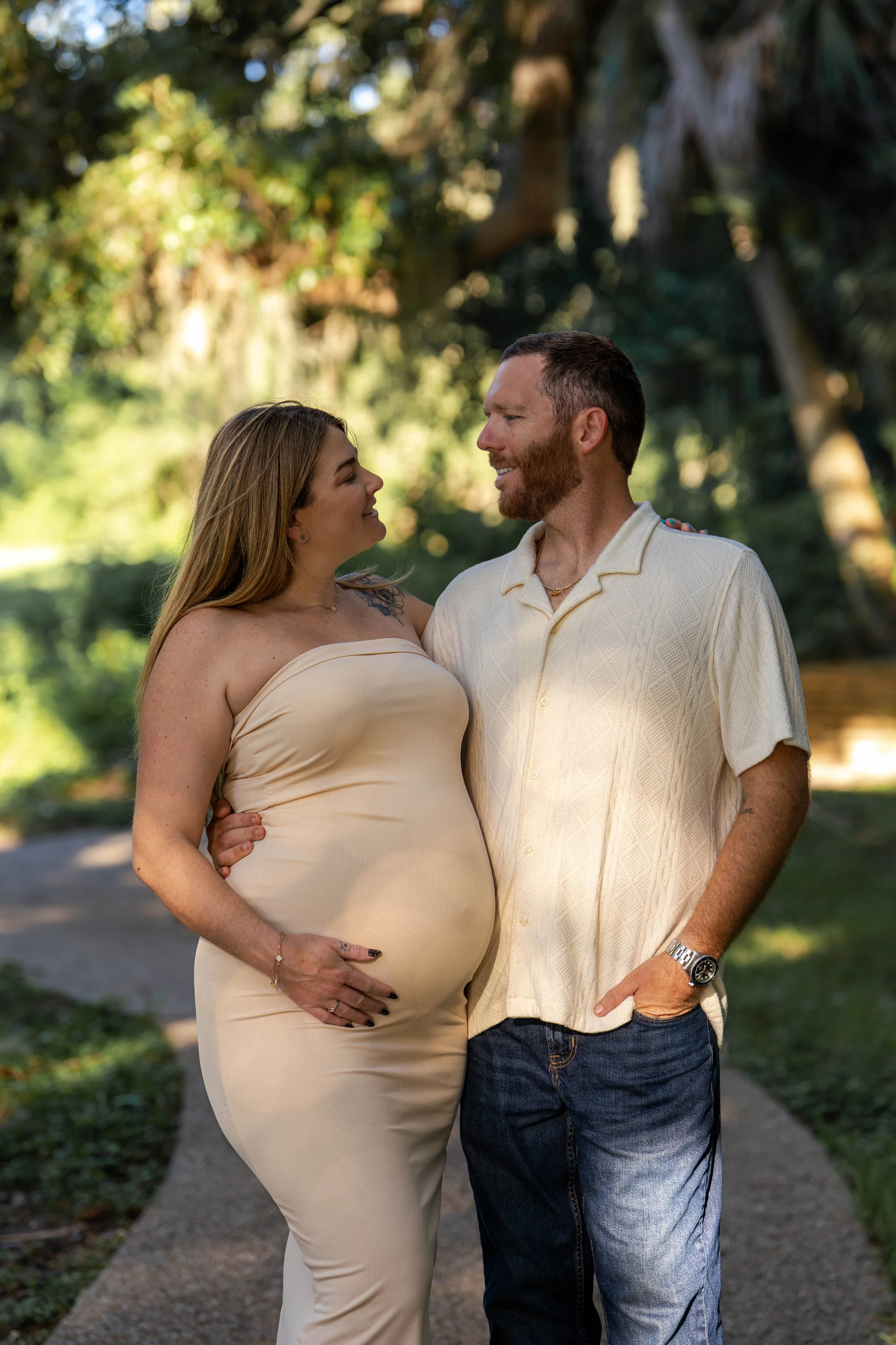 A pregnant woman and a man standing close together on a park pathway, smiling at each other, with trees and greenery in the background.