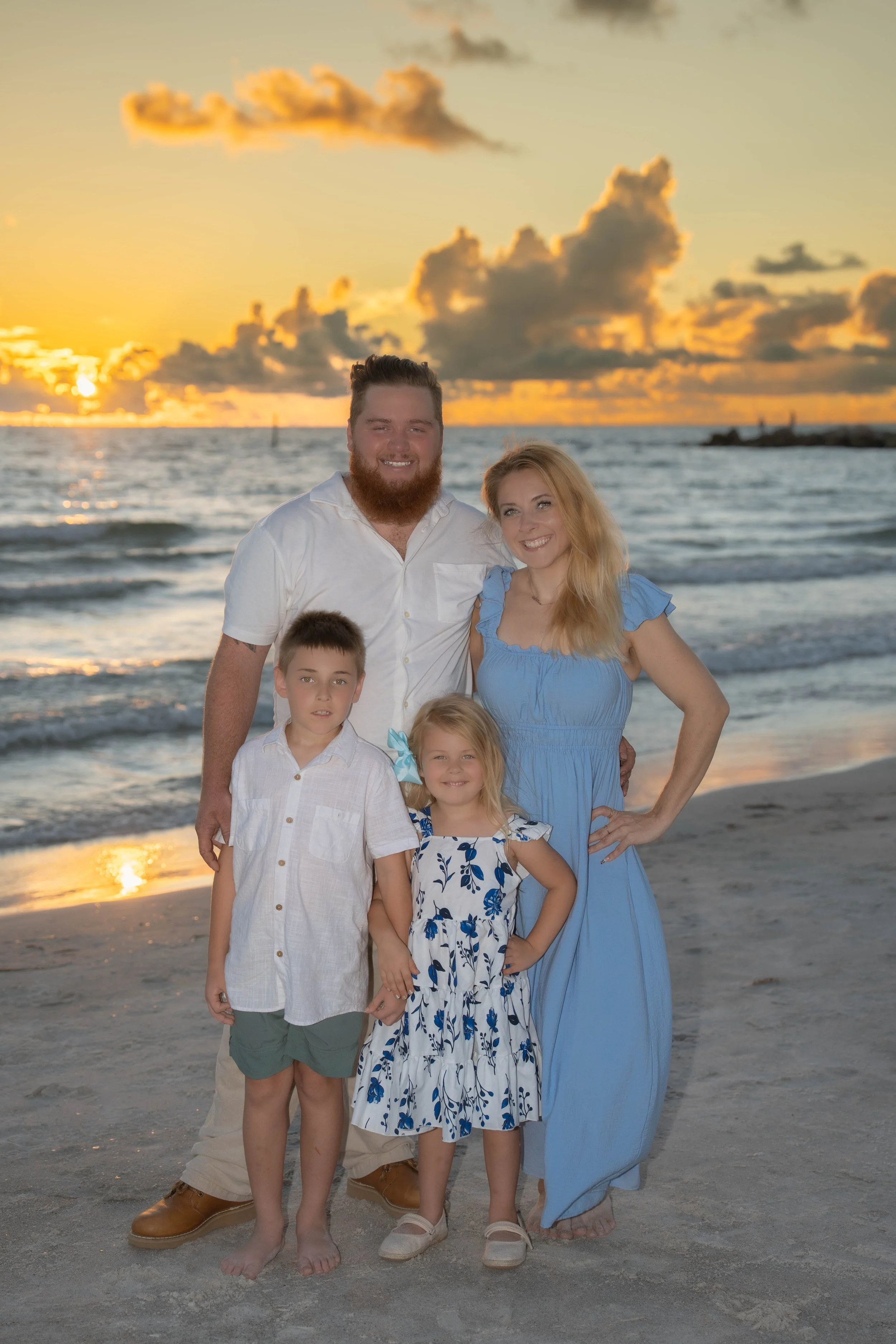 A family of six, including two adults and four children, standing on a beach during sunset with the ocean and a partly cloudy sky in the background.