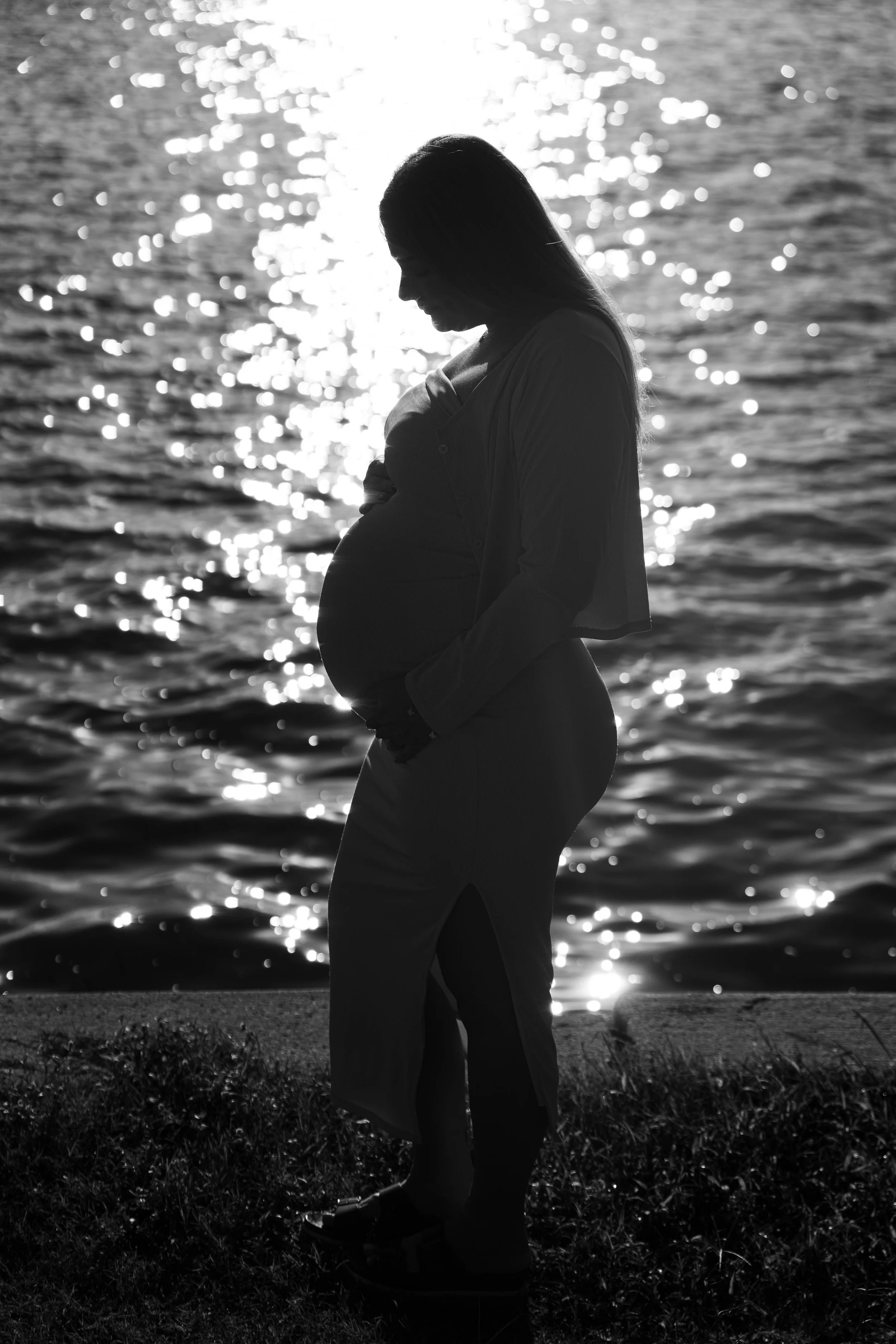Silhouette of a pregnant woman standing by a body of water with the sun reflecting on the surface, creating a bright, shimmering background.