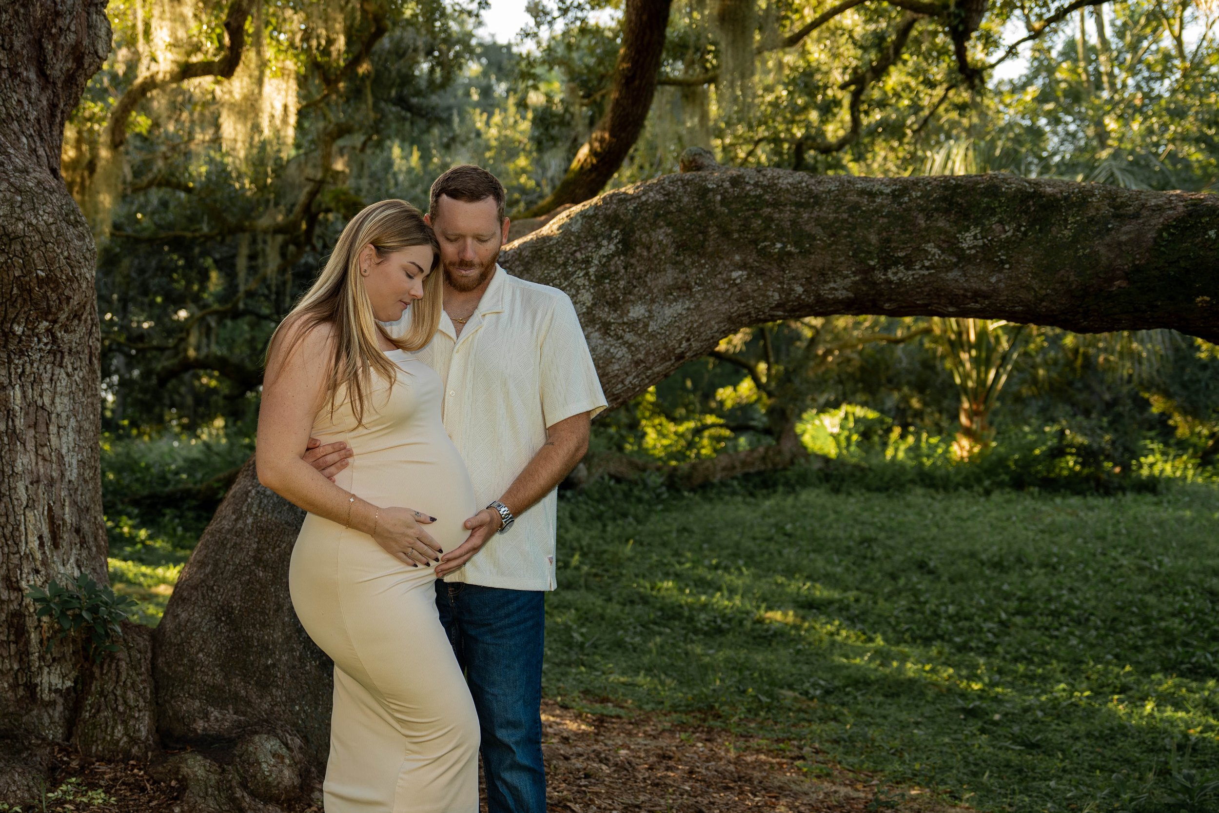 A pregnant woman and a man stand close together outdoors, with their foreheads touching and eyes closed, in a peaceful, wooded area with sunlight filtering through the leaves.