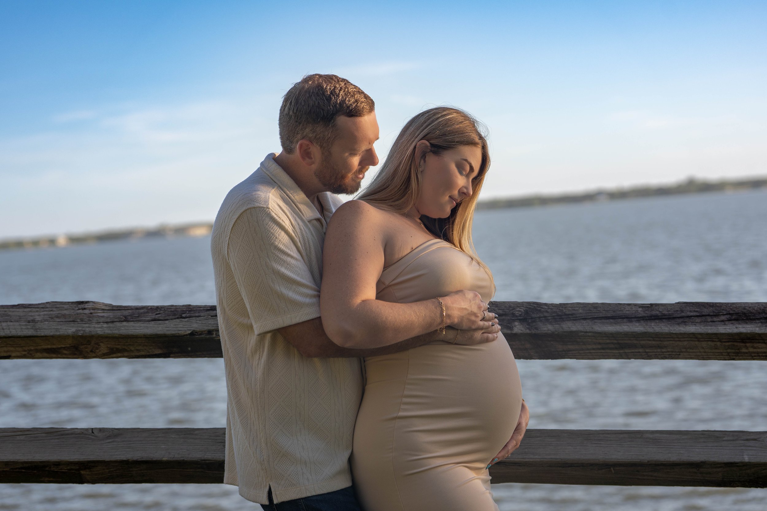 A pregnant woman and a man embracing next to a body of water, with the man holding her belly, both with eyes closed, during sunset.