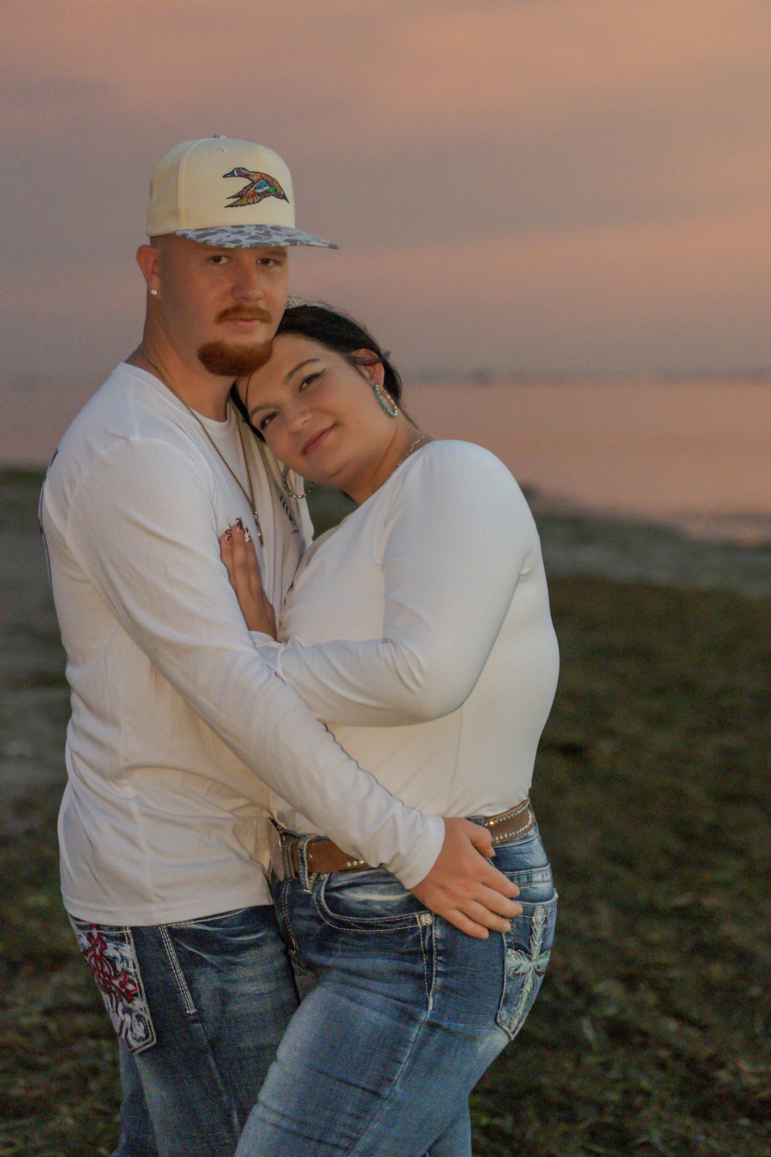 A couple standing closely together on a beach during sunset, embracing and smiling, with the ocean and sky in the background.