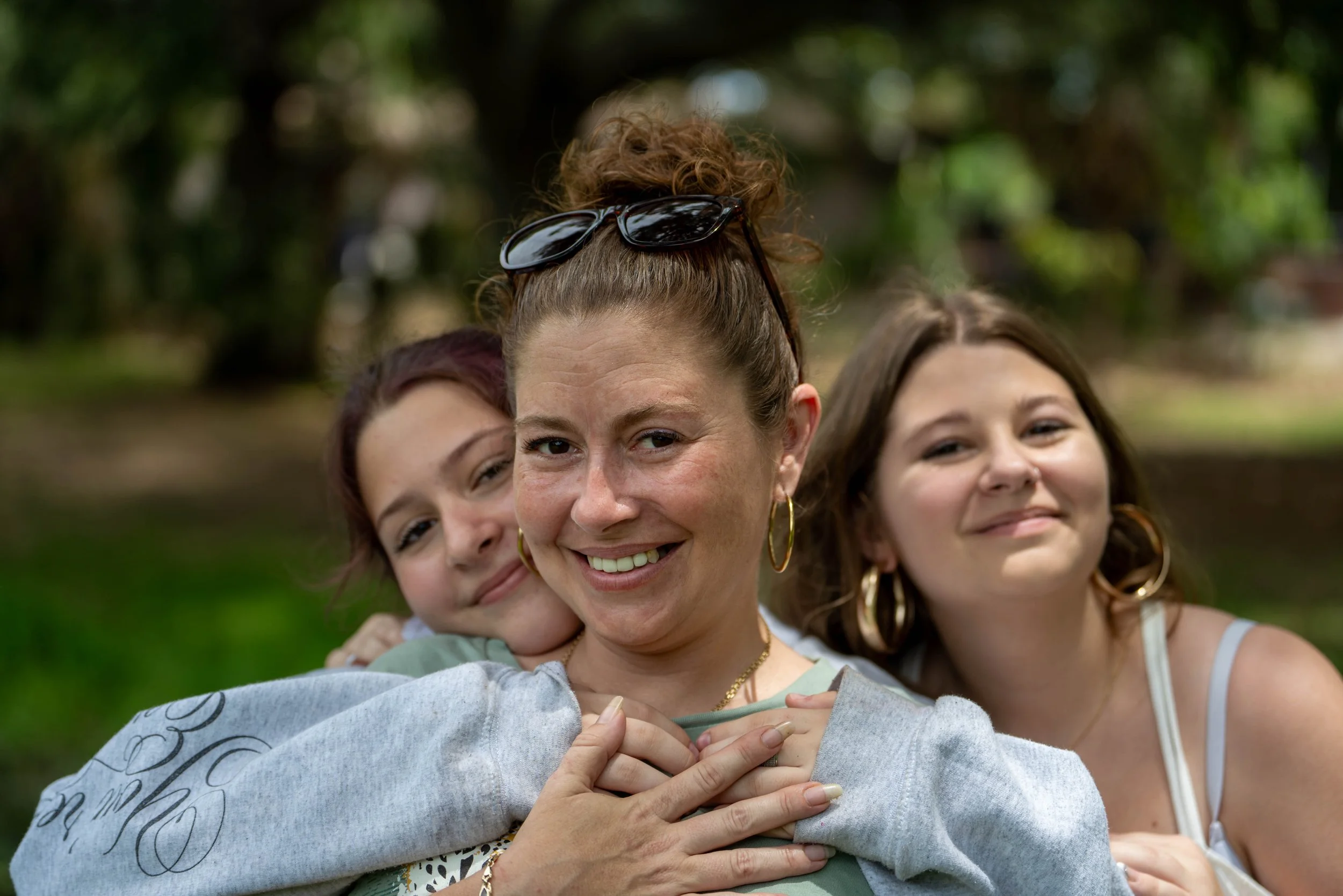 A woman with glasses on her head hugging two young girls outdoors, all smiling, in a park with trees in the background.
