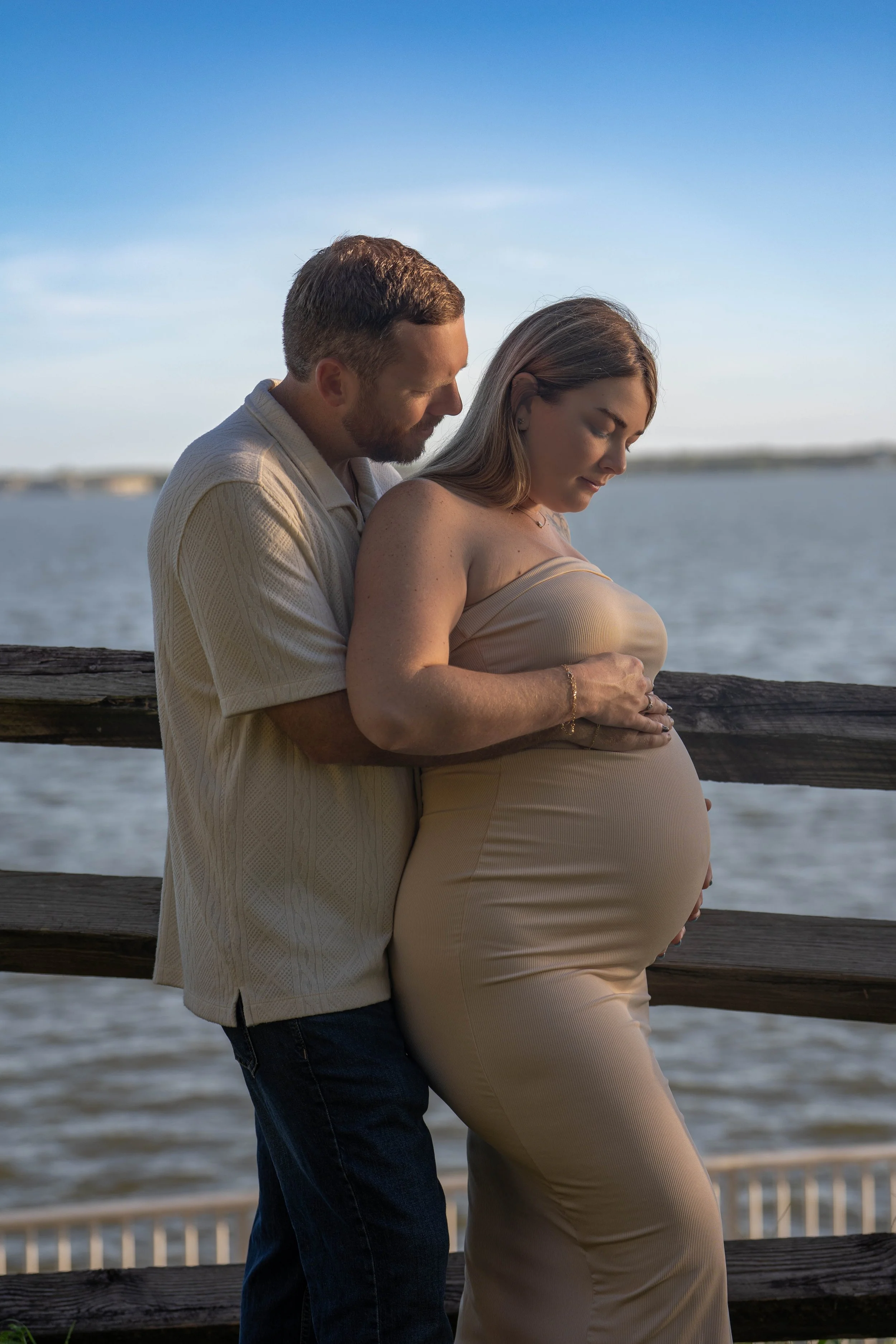 A man and pregnant woman standing together by a body of water, the man embracing the woman from behind with his head near hers, with a wooden railing in the foreground and a blue sky above.