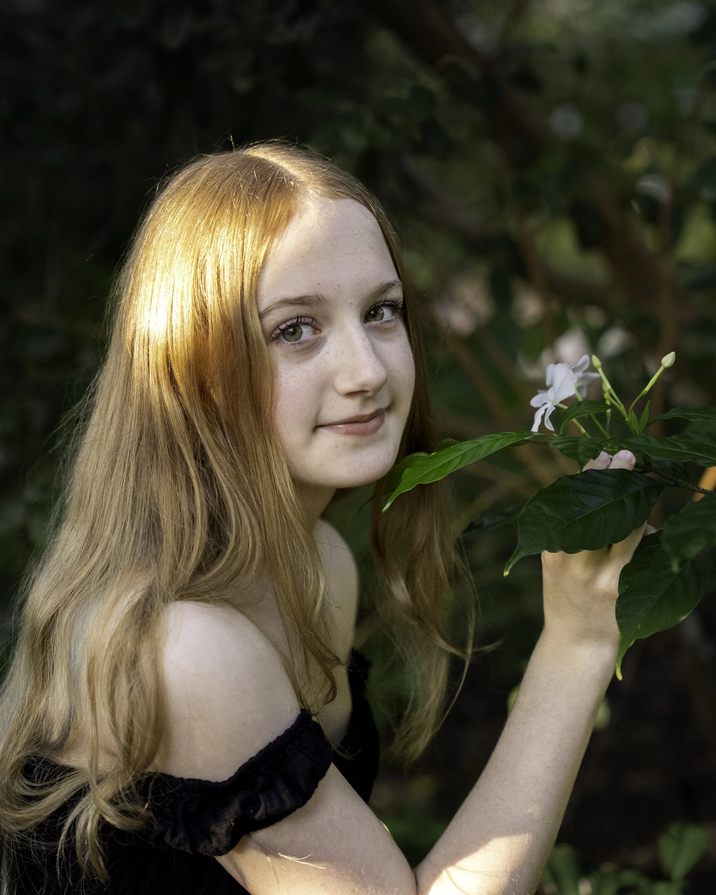 A young woman with long red hair and green eyes standing outdoors in a natural setting, holding a flowering plant with white flowers.