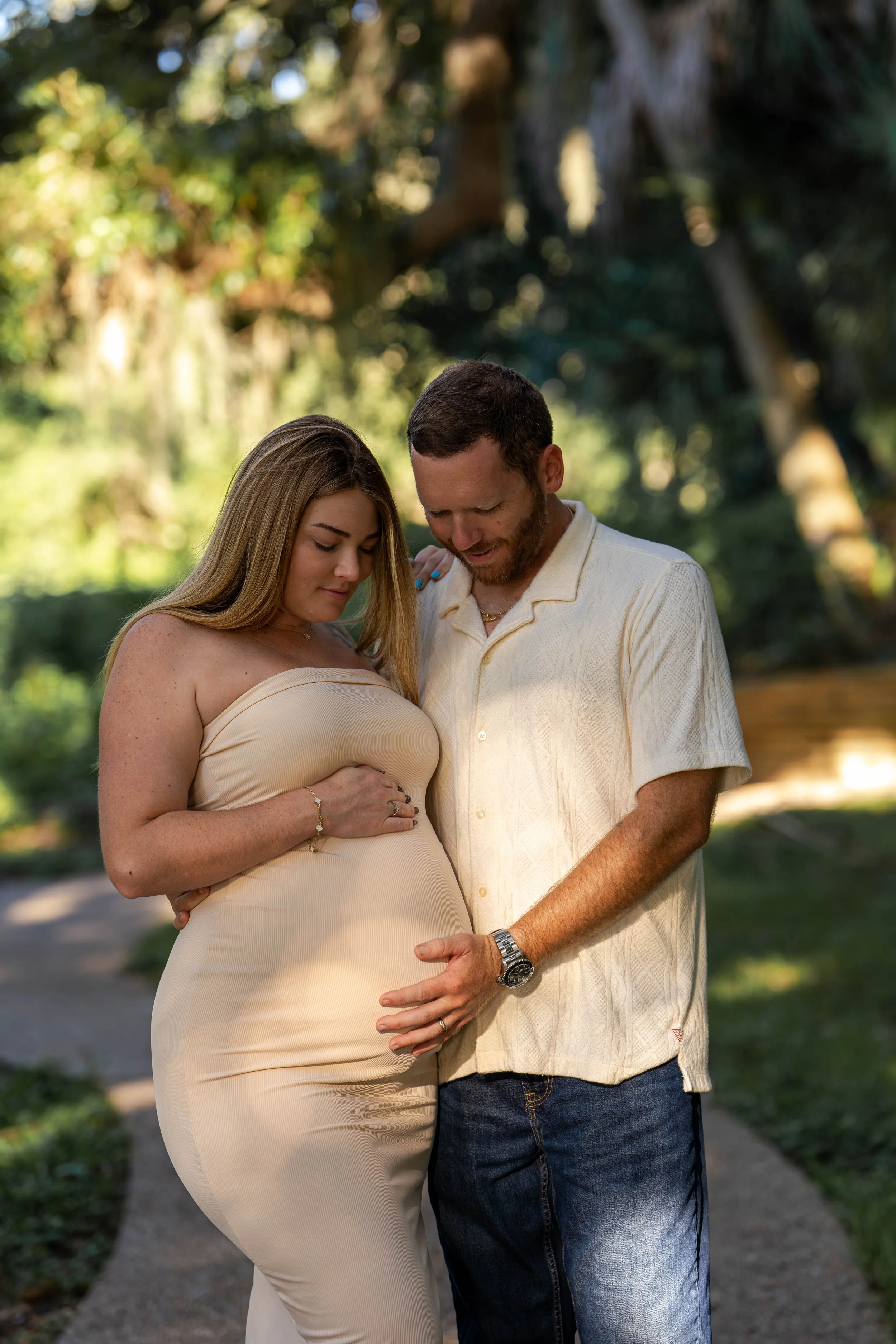 A pregnant woman in a beige dress with long hair and a man with a beard and short hair wearing a light-colored shirt and jeans, standing close together outdoors on a sunny day, with green trees and a pathway in the background.