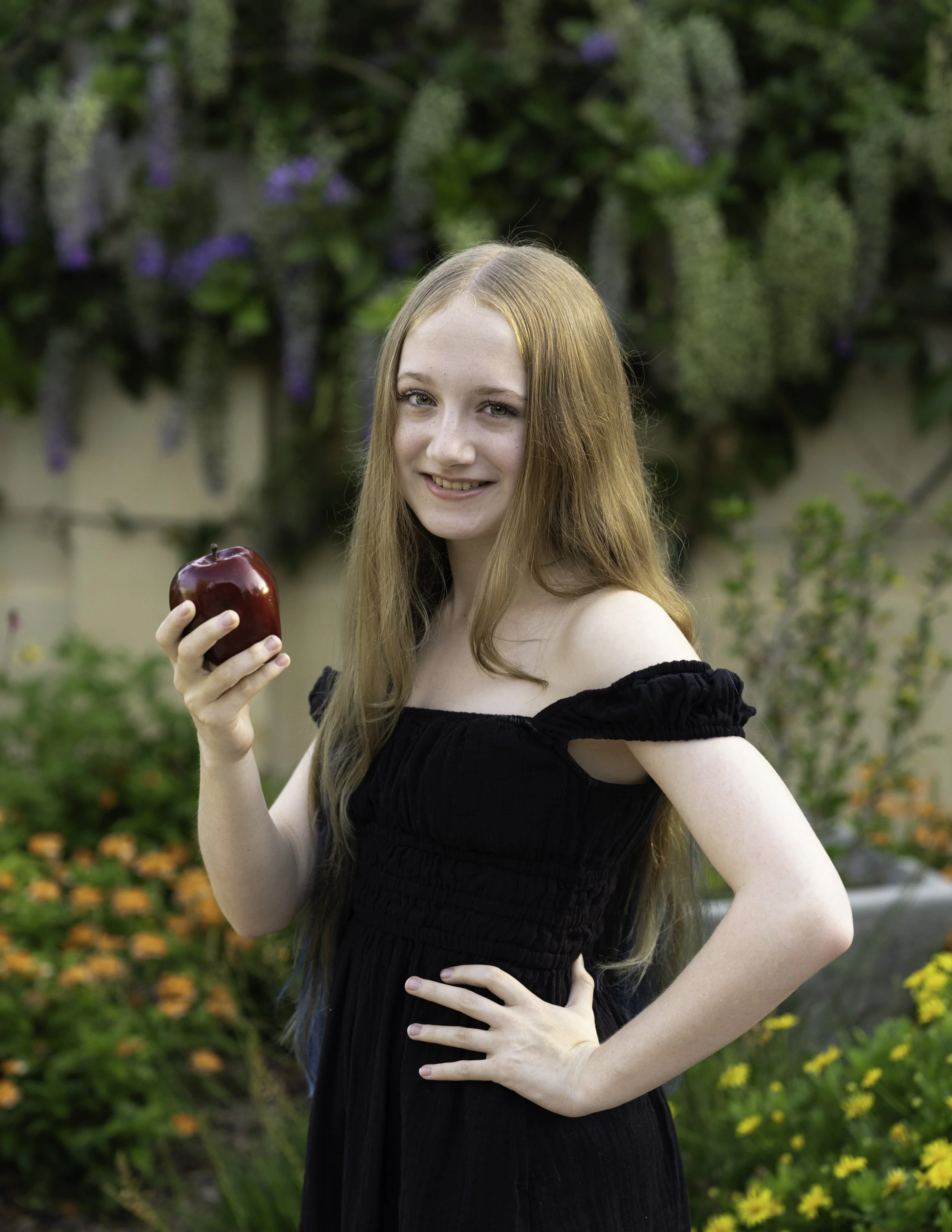 A young woman with long red hair, wearing a black off-the-shoulder dress, smiling and holding a shiny red apple in her right hand, standing outdoors in a garden with green foliage, purple flowers, and yellow flowers in the background.