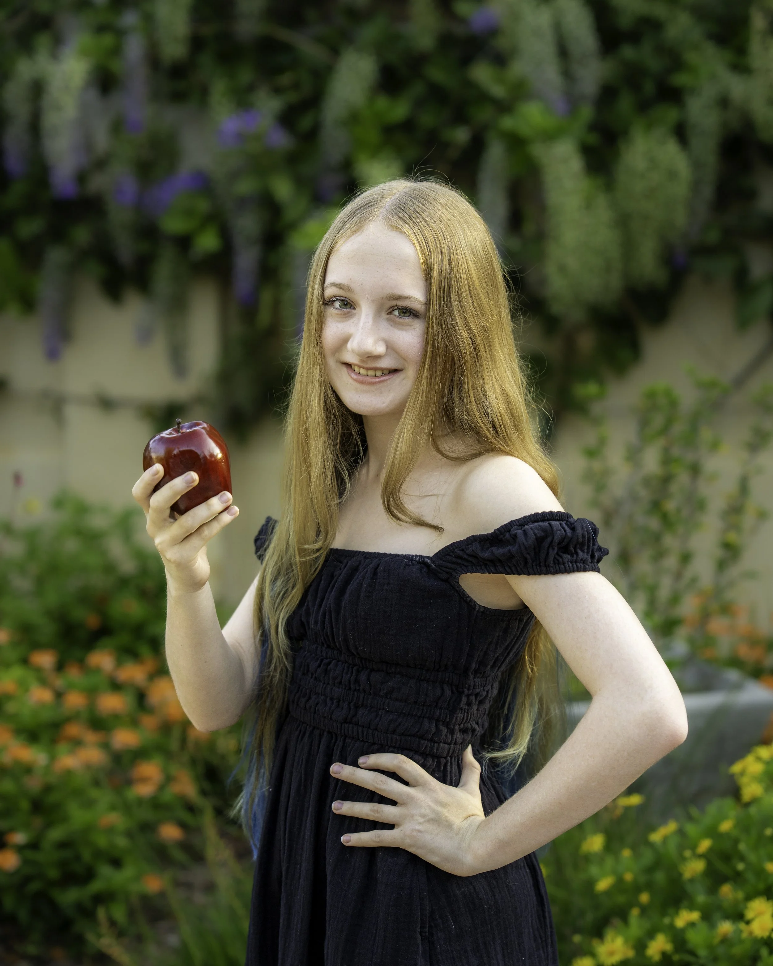 A young woman with long red hair smiling and holding a shiny red apple in an outdoor garden setting with green foliage and purple flowers in the background.