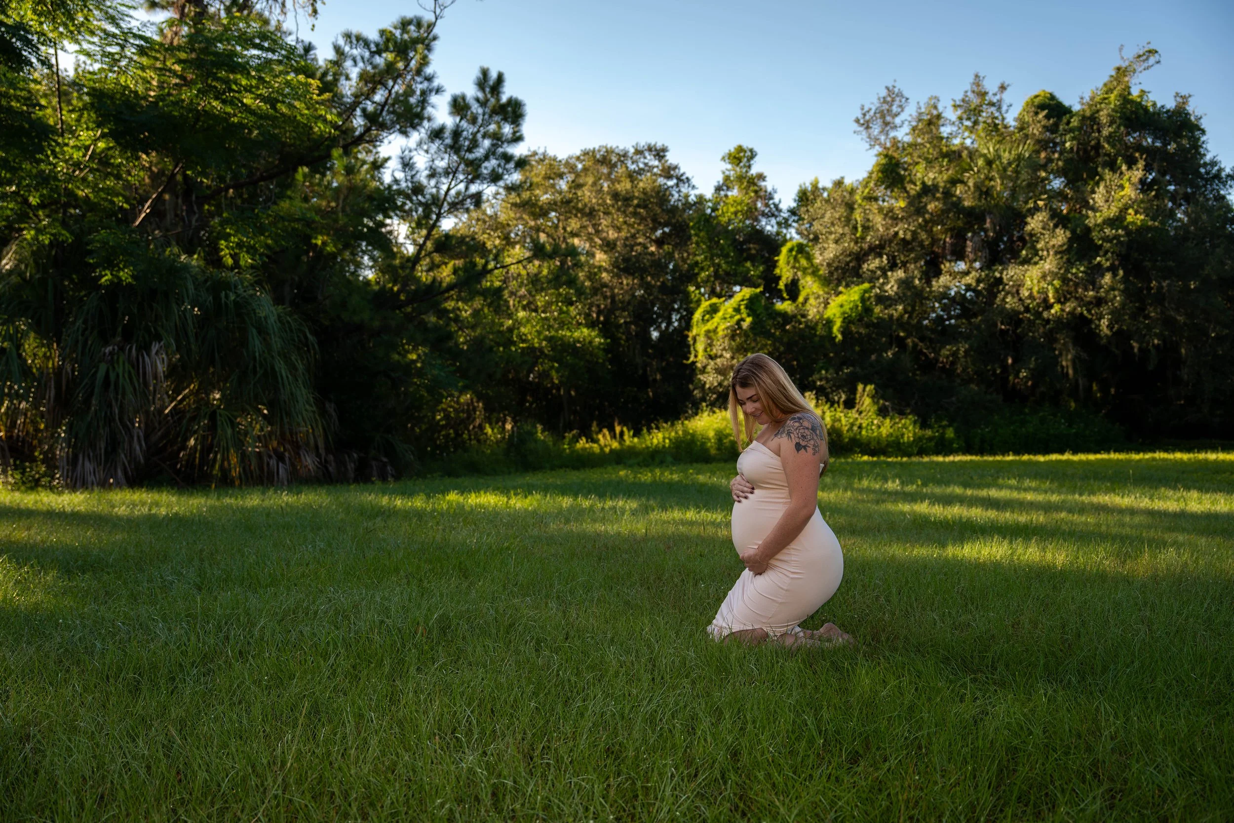 A pregnant woman in a beige dress kneeling on grass in a park, smiling, with her hands on her belly. She has shoulder-length blonde hair and a floral tattoo on her left shoulder. The park has lush green trees and clear blue sky.