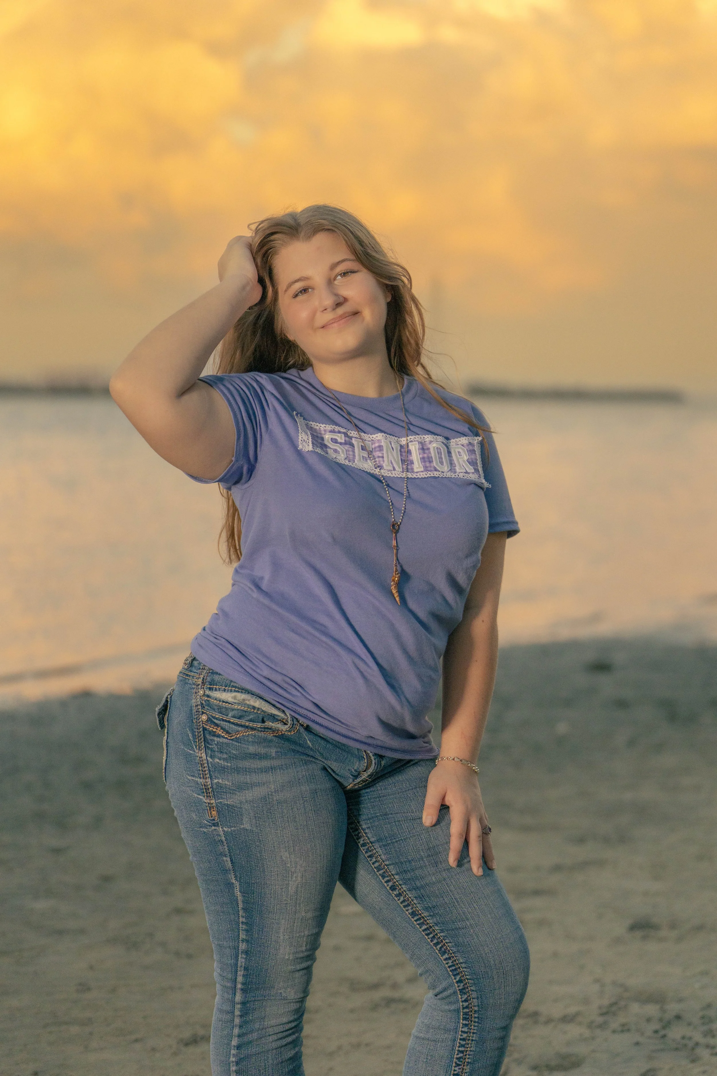 A young woman standing on the beach during sunset, wearing a purple t-shirt with 'Senior' written on it, denim jeans, and a necklace, smiling and touching her hair.