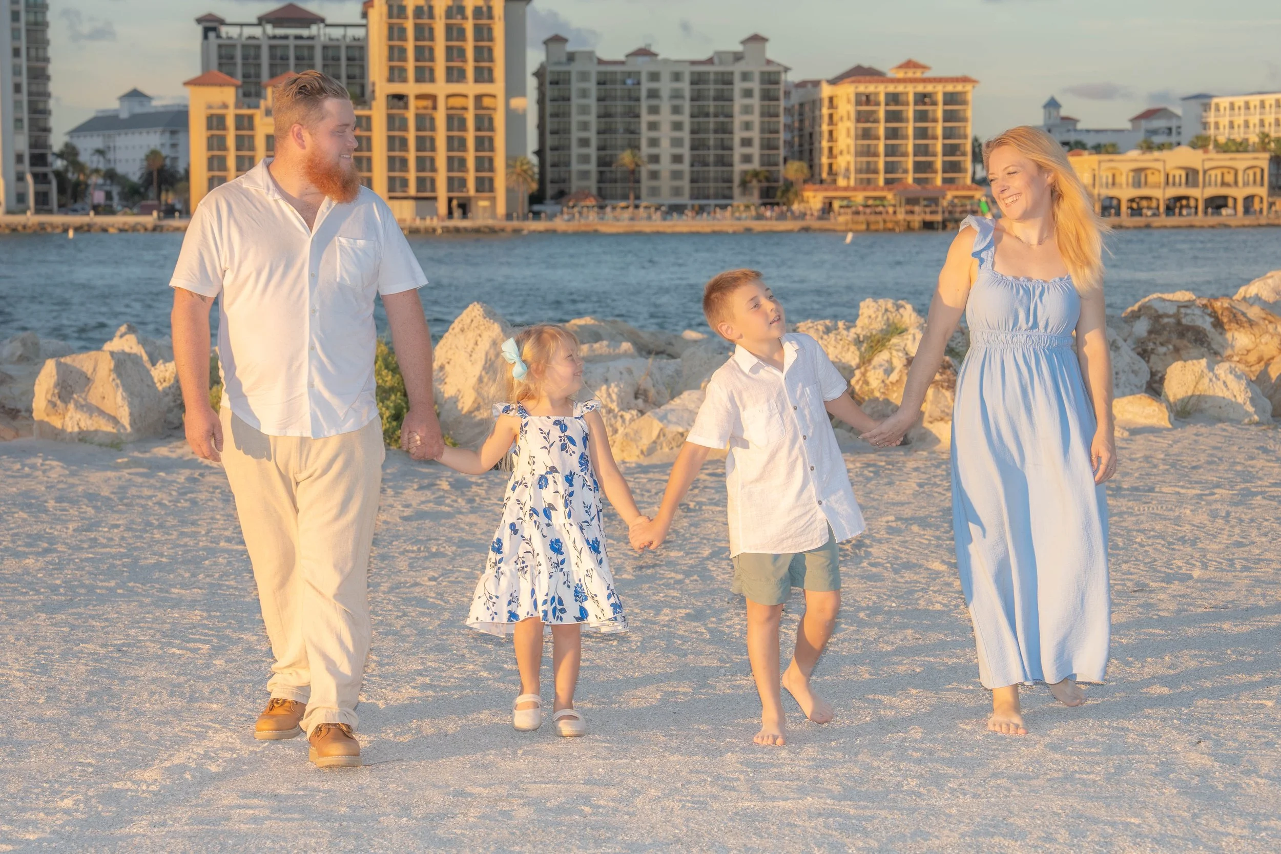A family holding hands and walking along a sandy beach at sunset with buildings and water in the background.