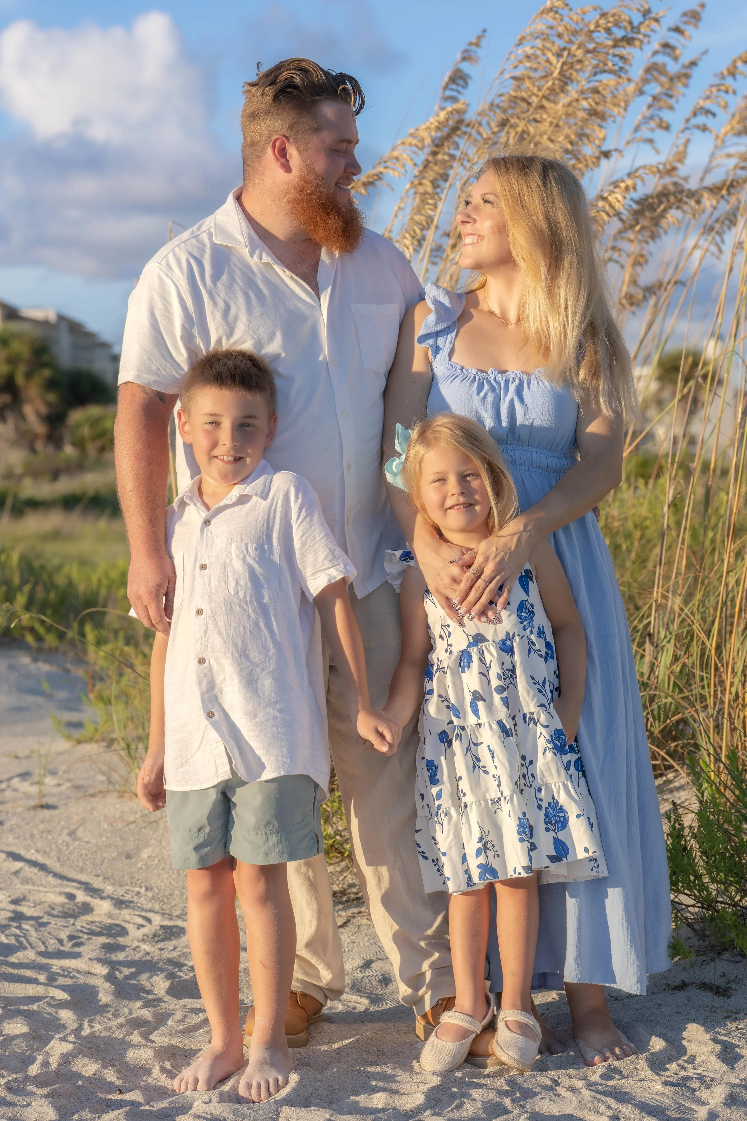 A family of four standing on a sandy beach with tall grass in the background. The father and mother are smiling at each other, holding their young daughter, while the son stands next to them. They are dressed in light summer clothing, enjoying a sunn