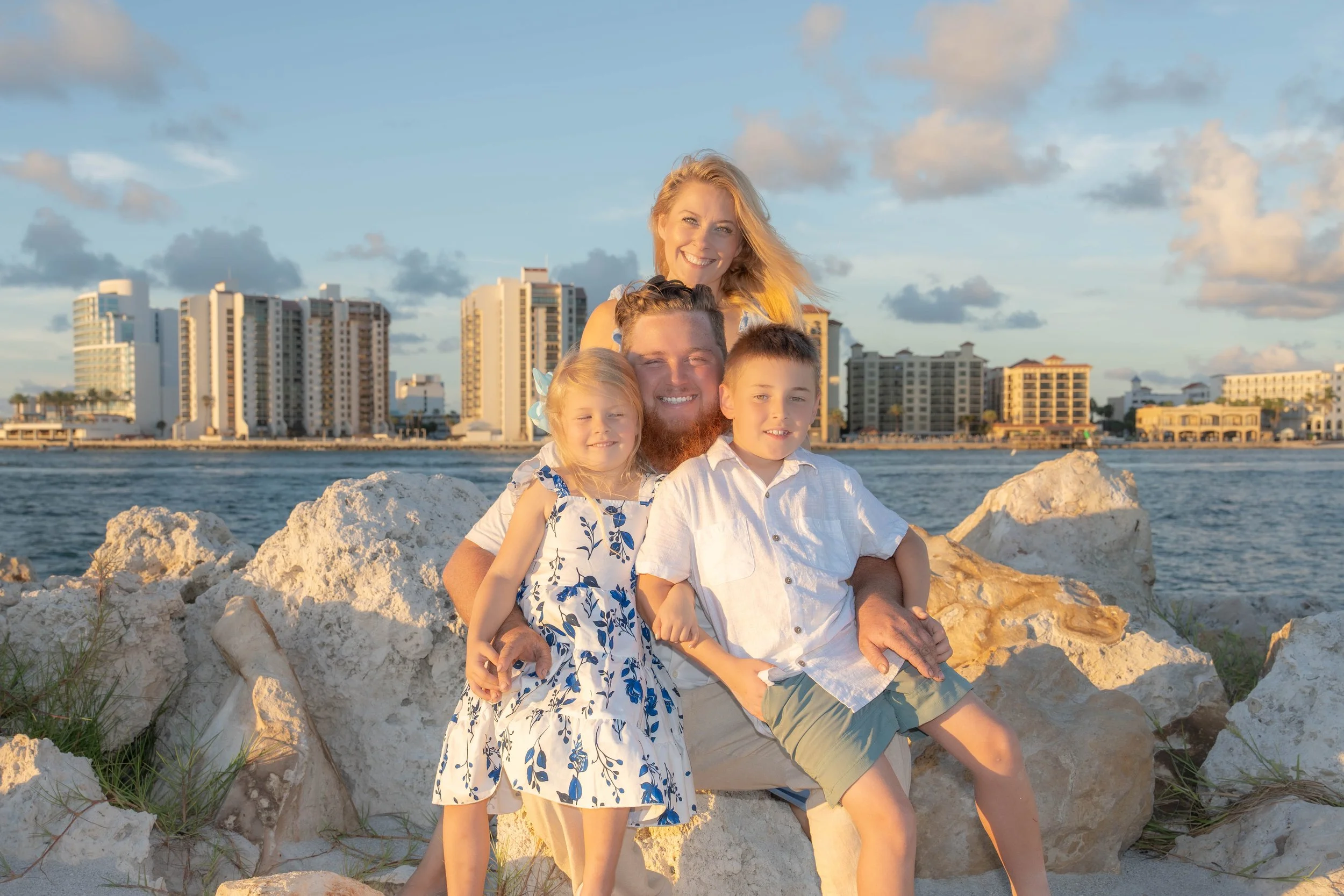 A family of five with blond hair sitting on rocks by the water with a cityscape of tall buildings in the background, during sunset.
