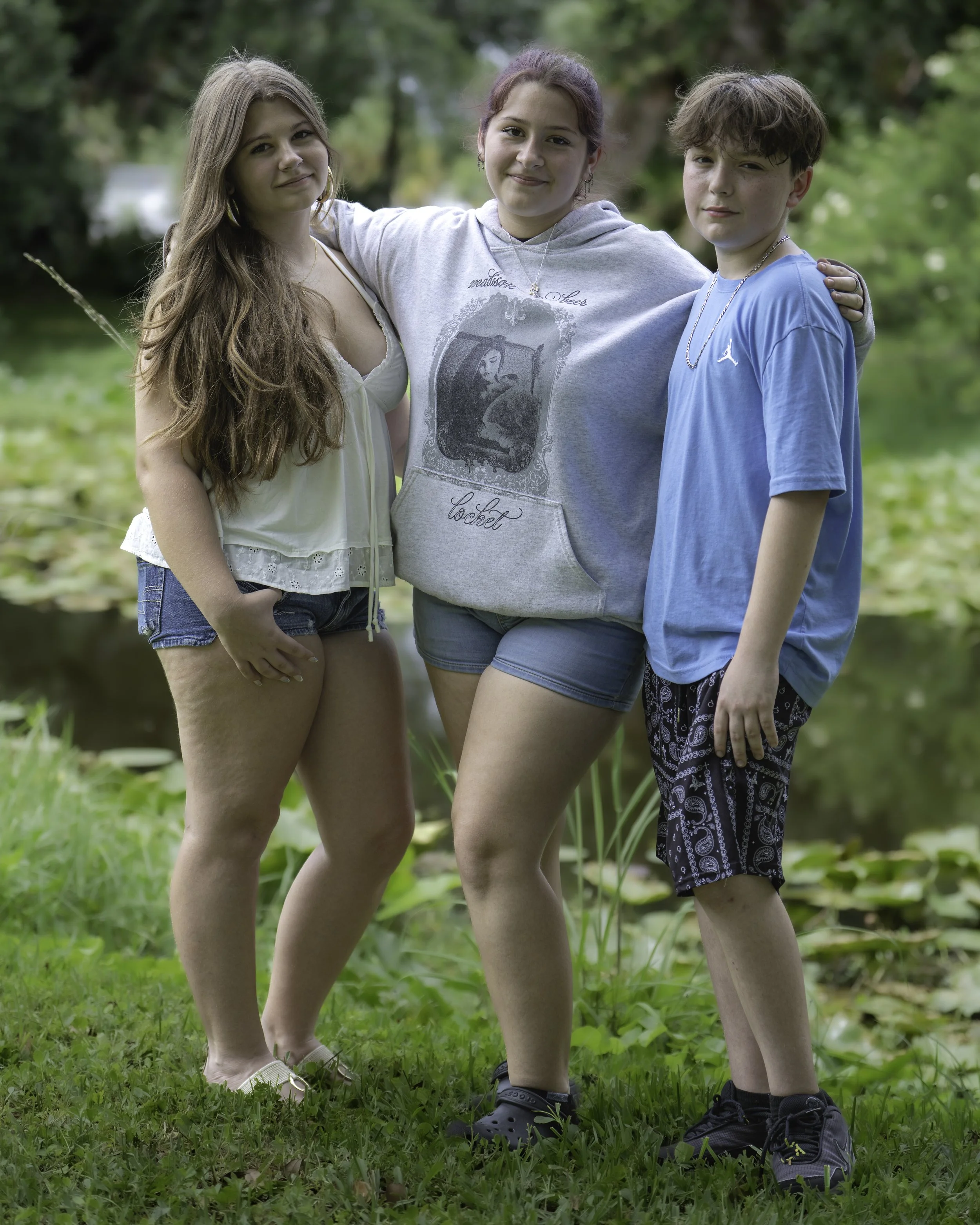 Three teenagers standing outdoors by a pond with green foliage in the background, smiling and posing for the photo. They are wearing casual summer clothes and sneakers.