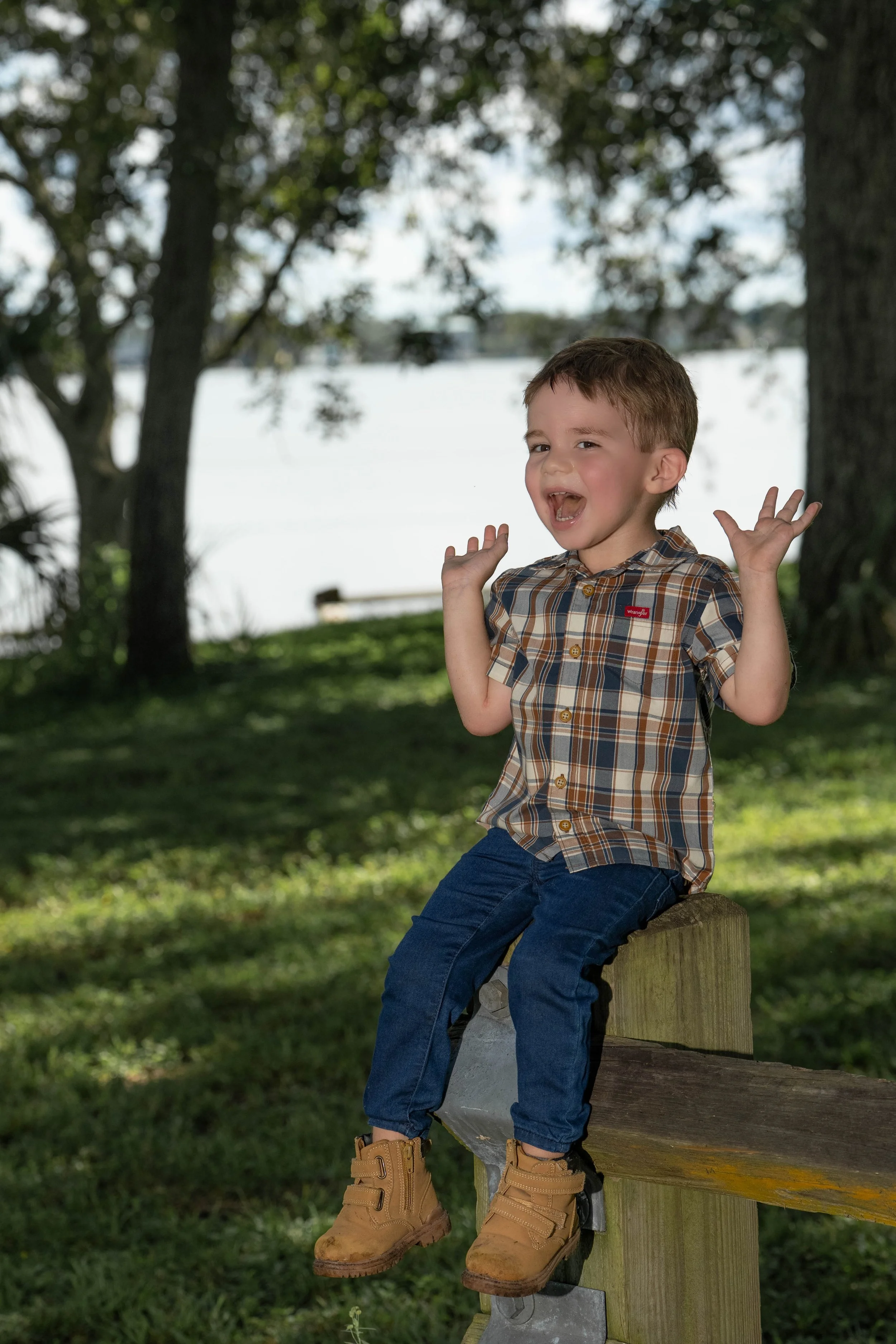 A young boy sitting on a wooden bench outdoors, surrounded by trees, smiling and raising his hands.