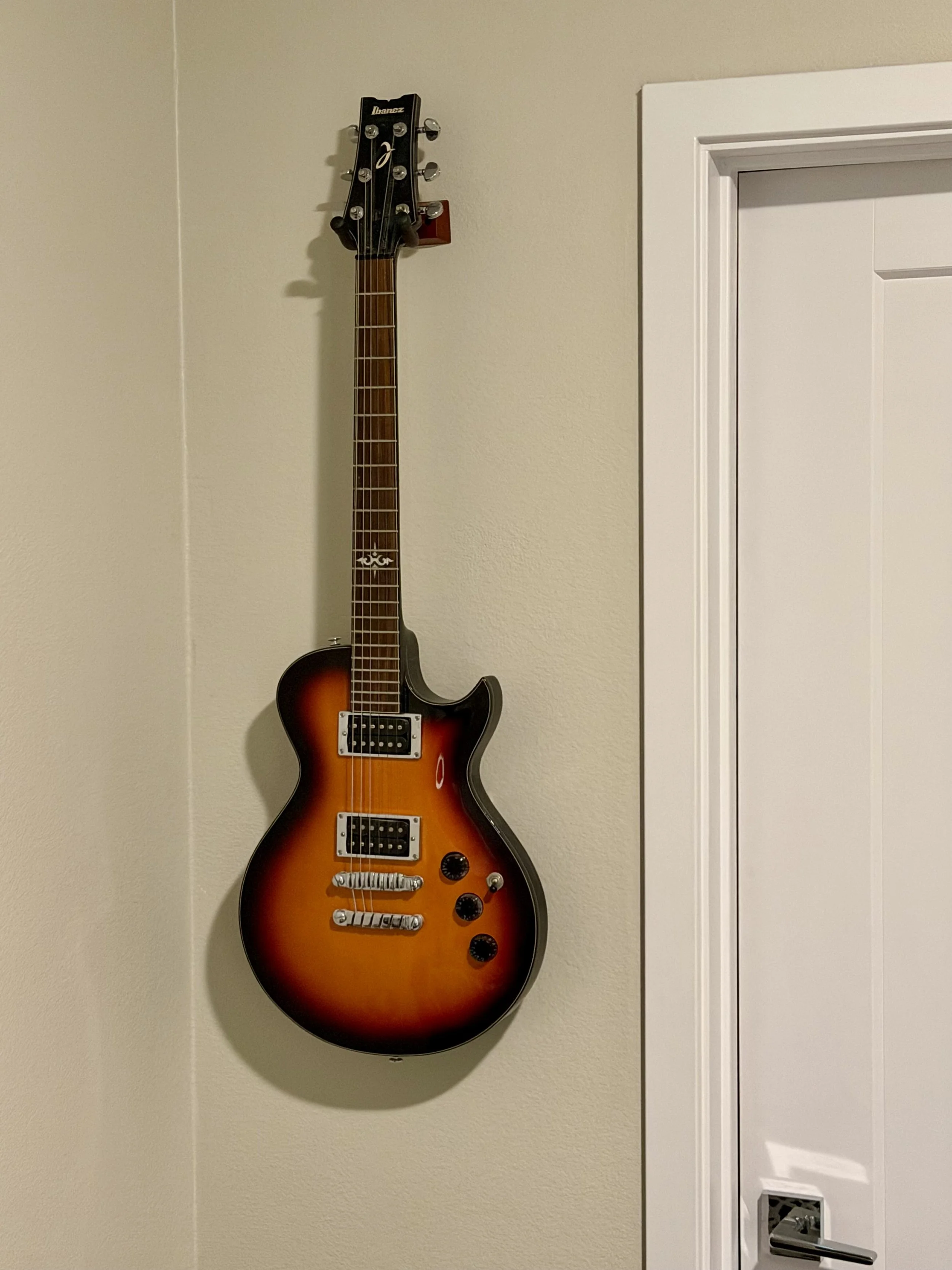 An electric guitar with a sunburst finish hanging on a beige wall, next to a white door with a silver handle.