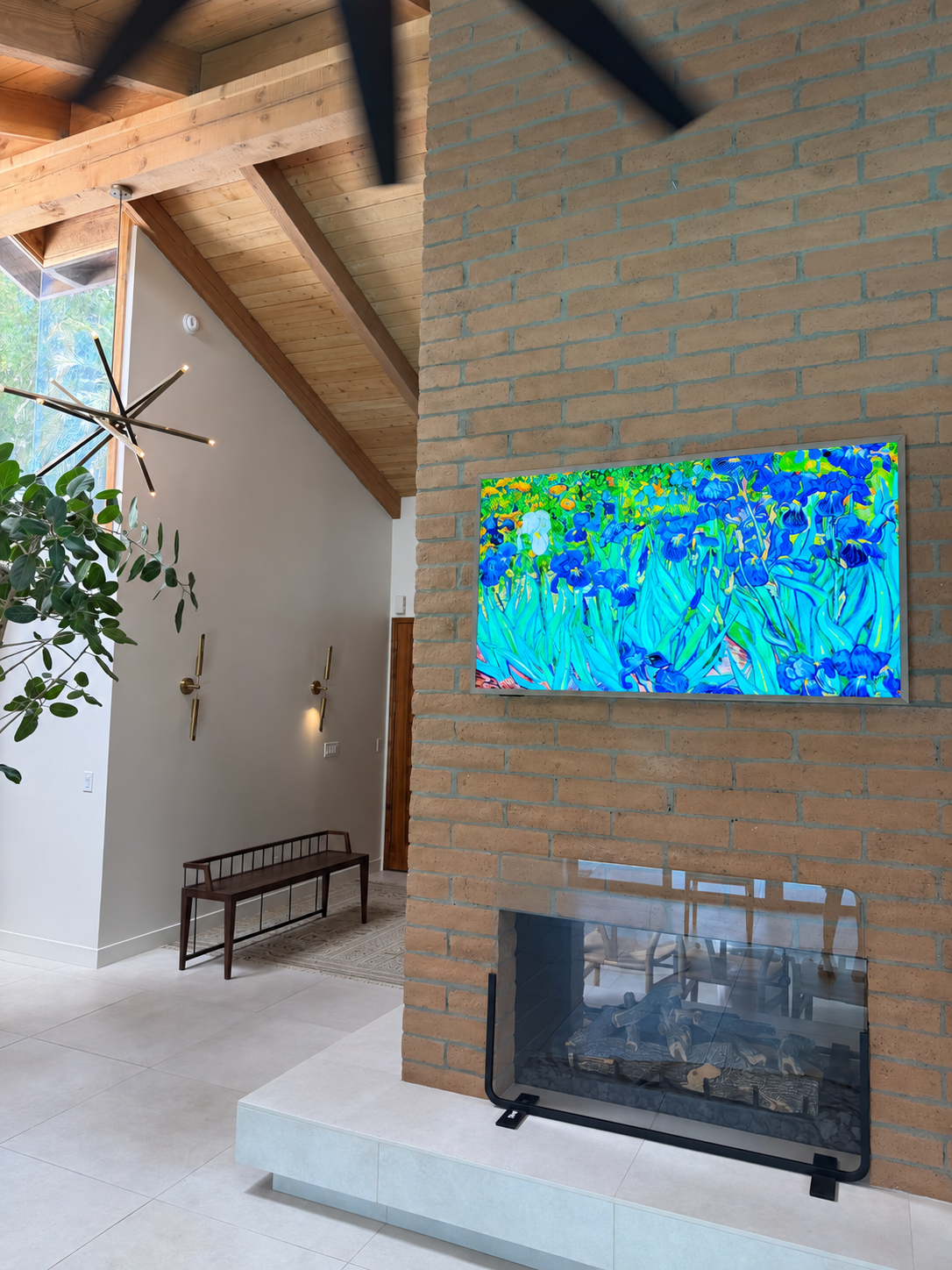 Modern living room with brick fireplace, mounted TV displaying colorful abstract art, and a wood and metal bench beneath brass wall sconces. A large green plant and contemporary chandelier are also visible.