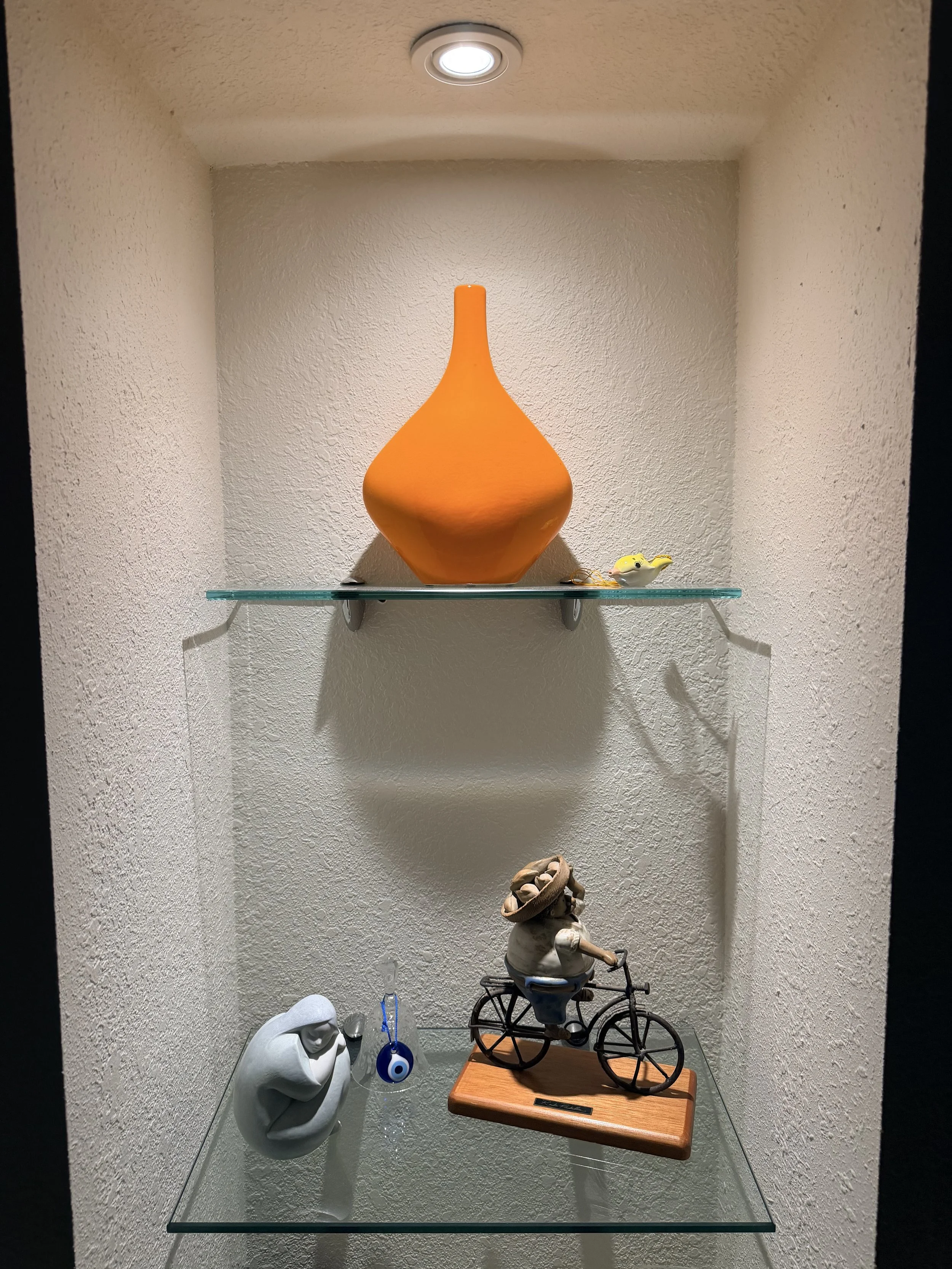 Display of decorative objects on glass shelves, including an orange vase, a small bird figurine, a white decorative sculpture, a keychain with a blue eye, and a metal sculpture of a person riding a bicycle, set against a textured beige wall.