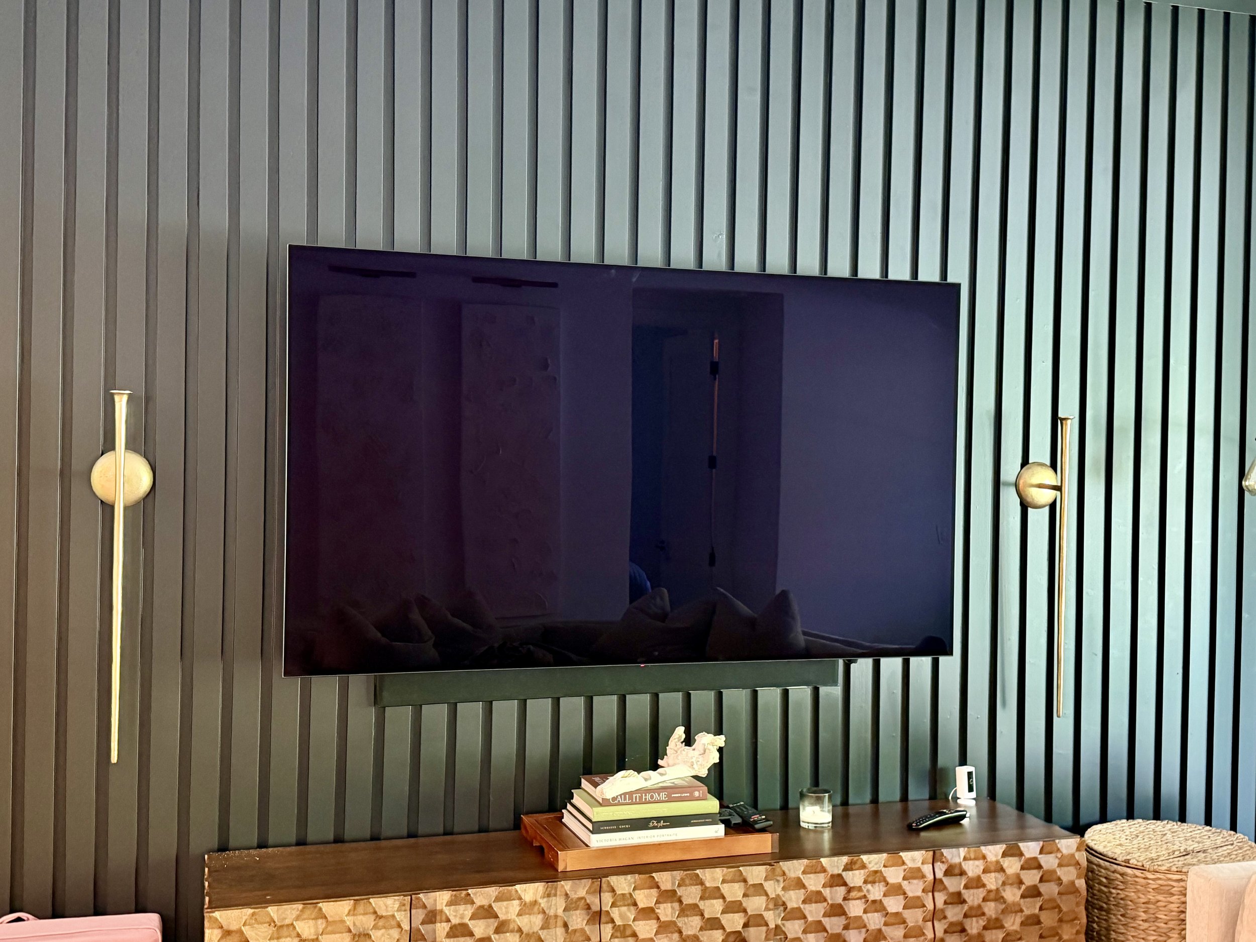 Living room wall with vertical gray paneling, mounted flat-screen TV, wooden side table with stacked books, and decorative objects.