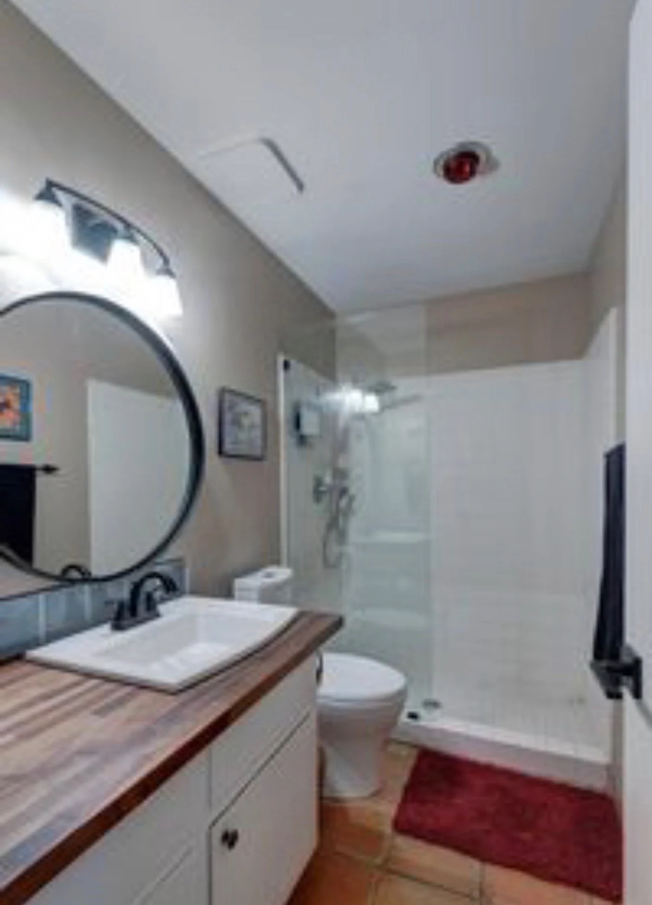 A bathroom with a wooden countertop sink, mirror, white toilet, shower with glass door, red rug, and walls decorated with framed pictures.