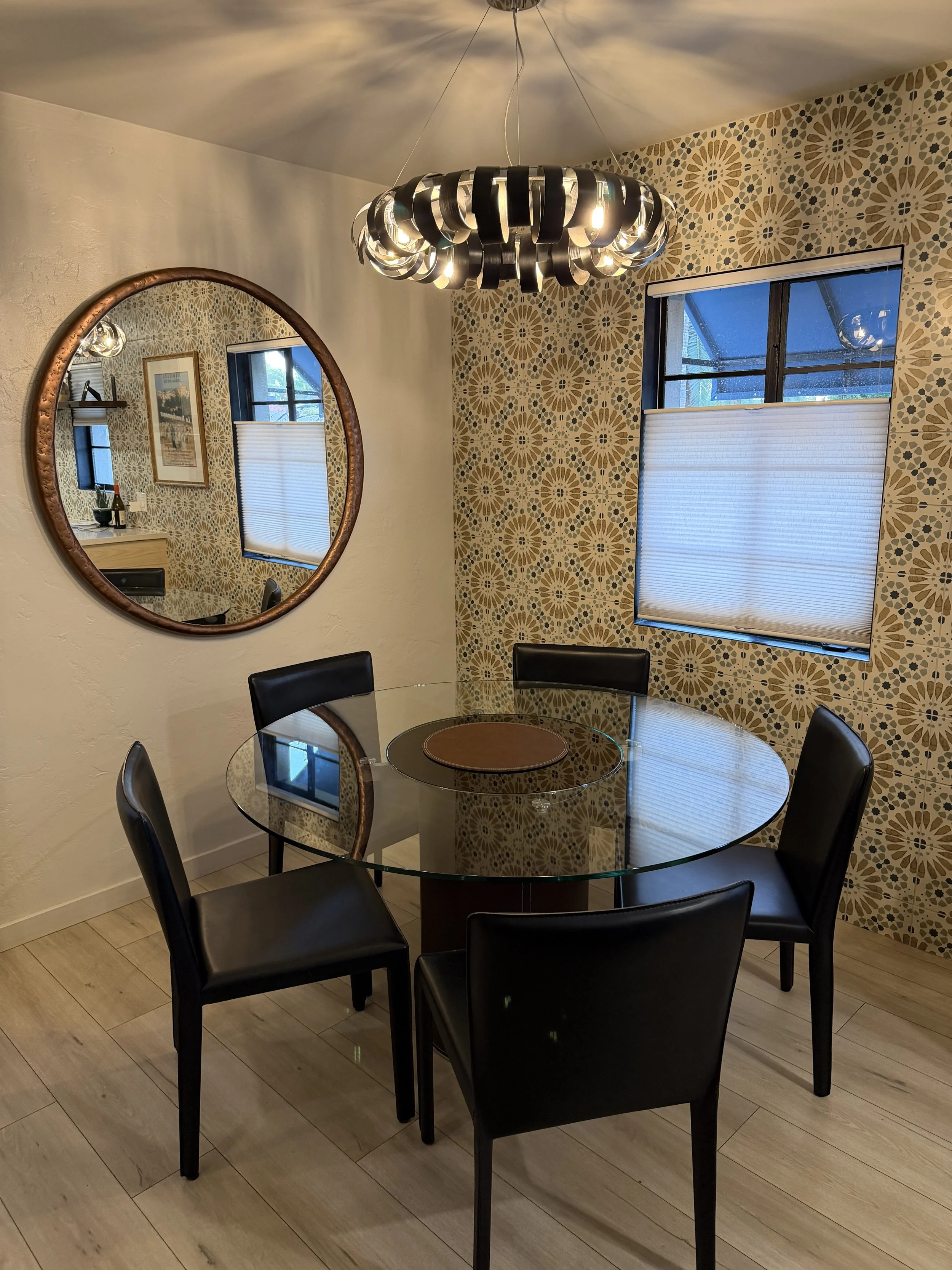 A dining room with a round glass table surrounded by five black chairs with cushioned seats. There's a large, modern black chandelier with circular elements hanging from the ceiling. On the wall, there's a decorative wallpaper with a geometric patter