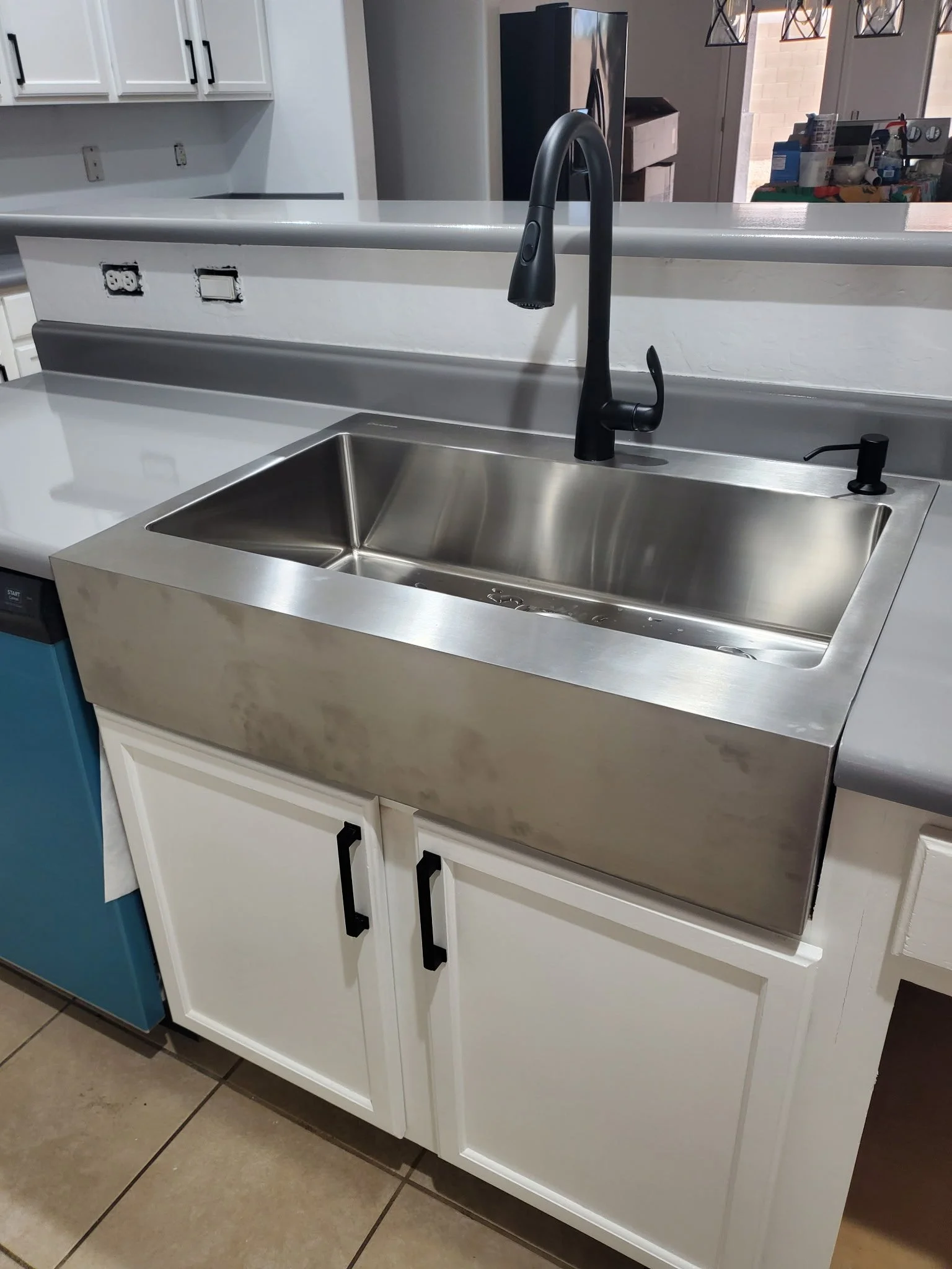 A kitchen sink with a black faucet on a white counter. There are upper white cabinets and outlets missing covers. The background shows a refrigerator and some sealed boxes.
