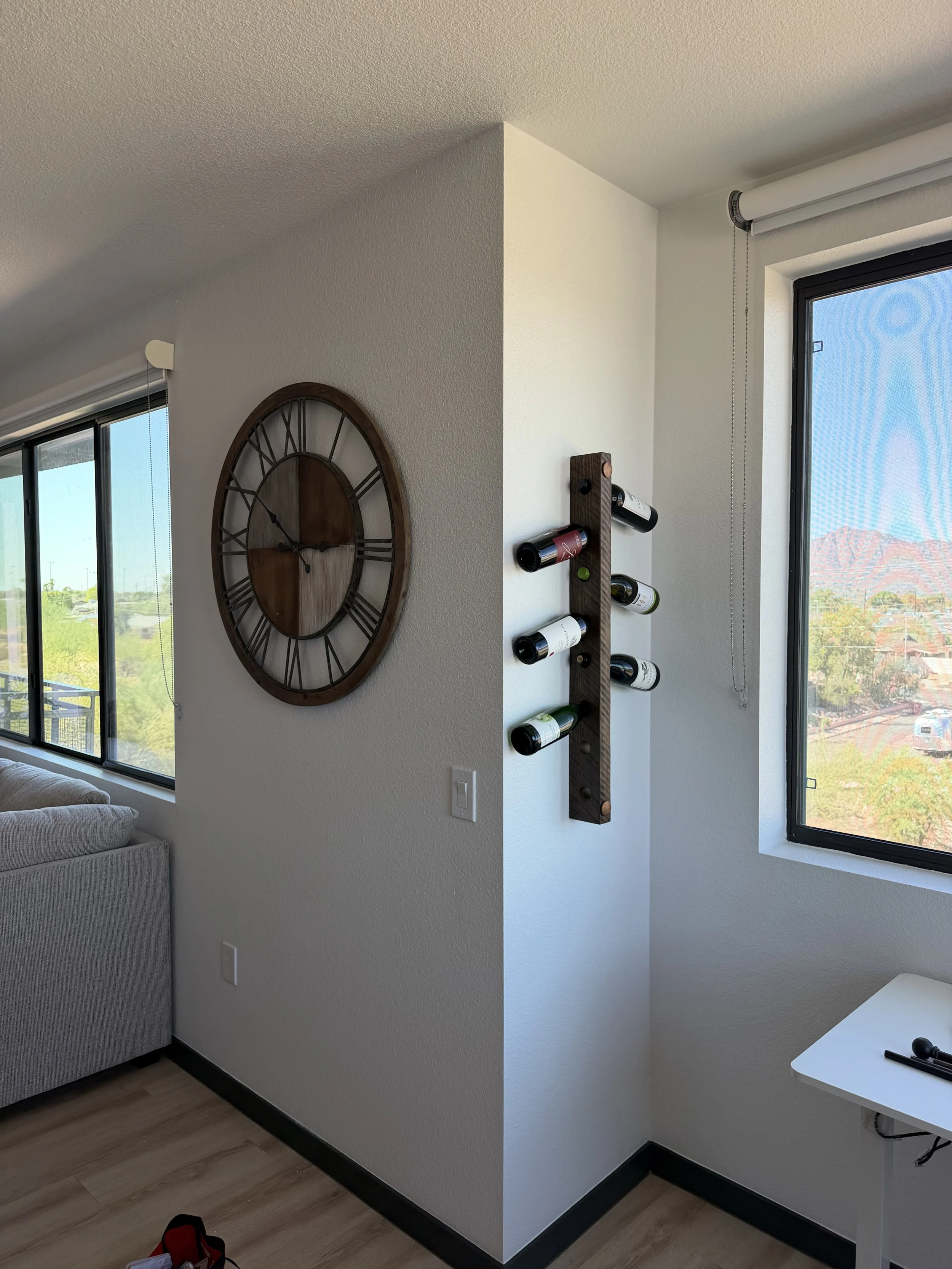 Corner of a bright room with a large wall clock and a wine rack on the wall, two large windows with blinds showing a mountain landscape and clear sky, part of a beige sofa, and a small white table with a remote control and pen.