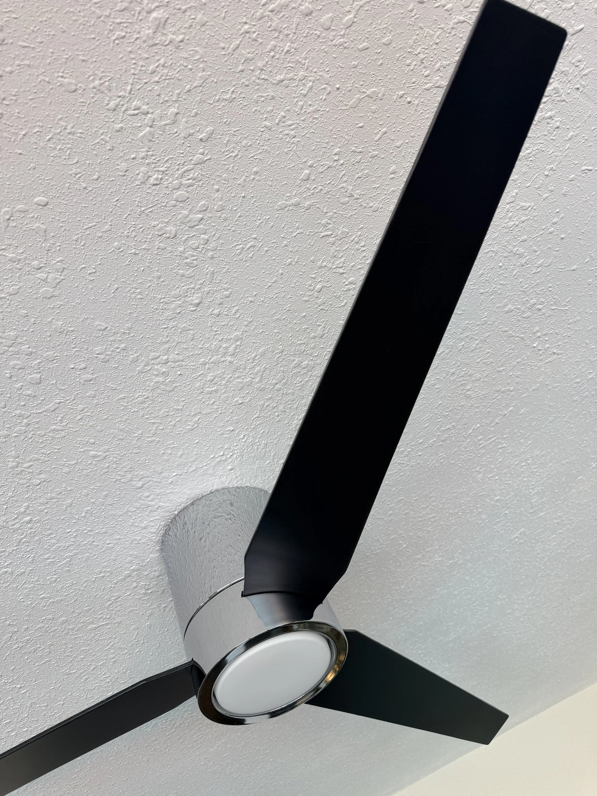 A black ceiling fan with three blades and a round white light fixture mounted on a white textured ceiling.