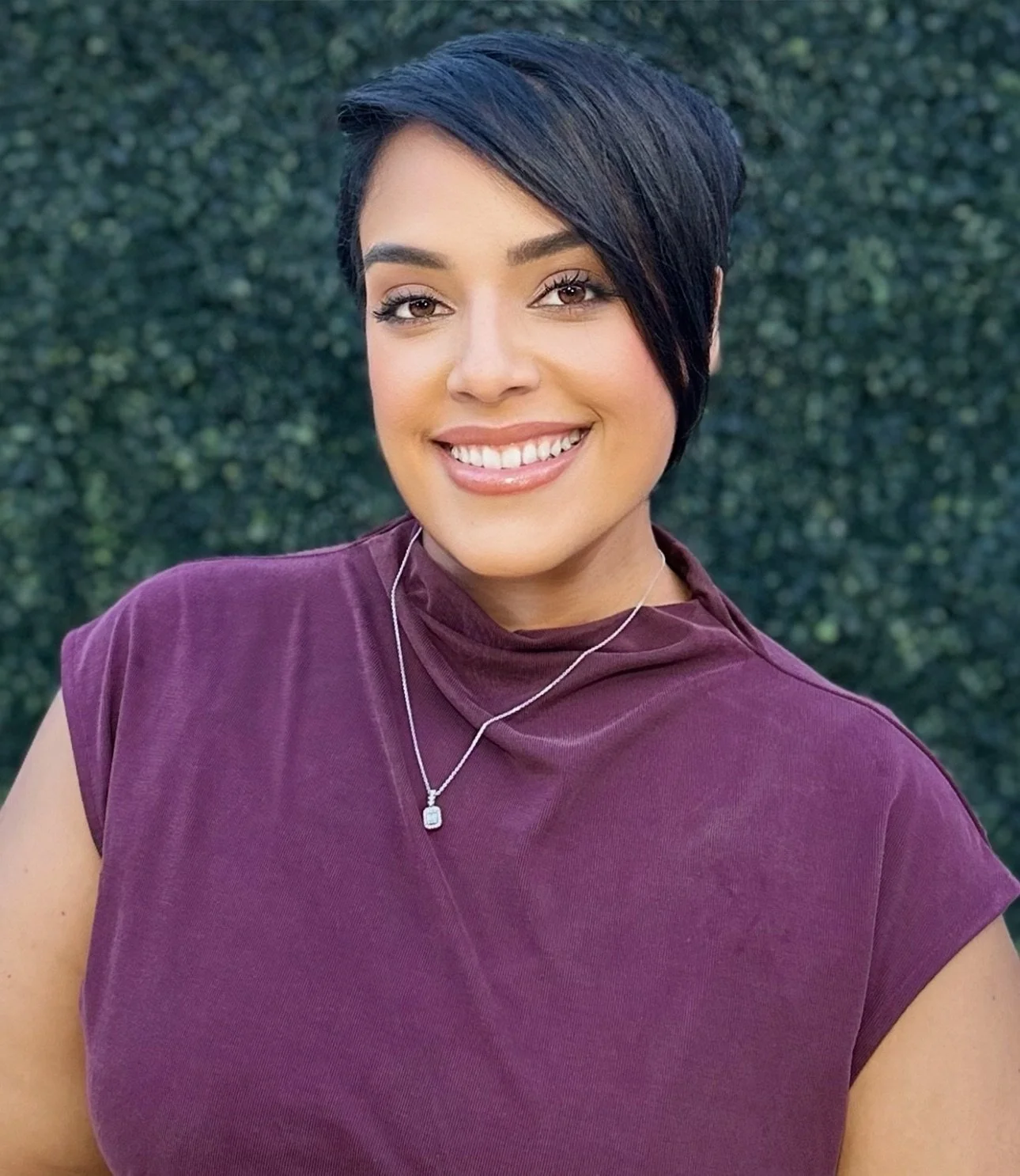 A woman with short black hair, wearing a purple top and silver necklace, smiling outdoors against a hedge background.