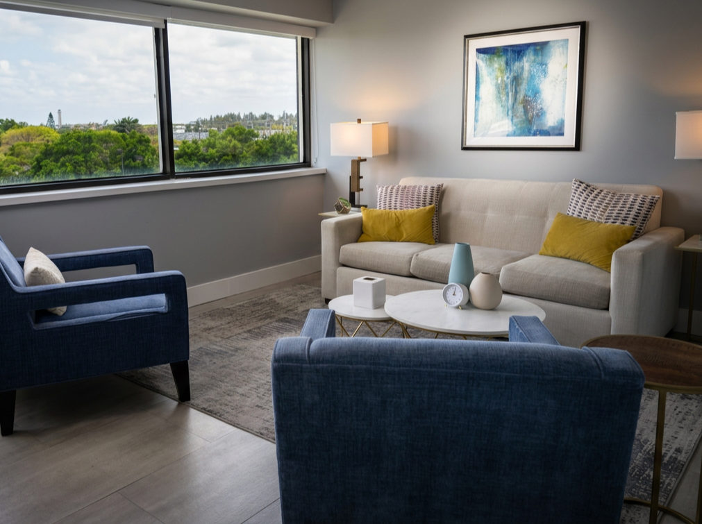 Modern living room with a beige sofa, two blue armchairs, and a white coffee table decorated with vases and clocks, large window with a city view, abstract wall art, and table lamps.