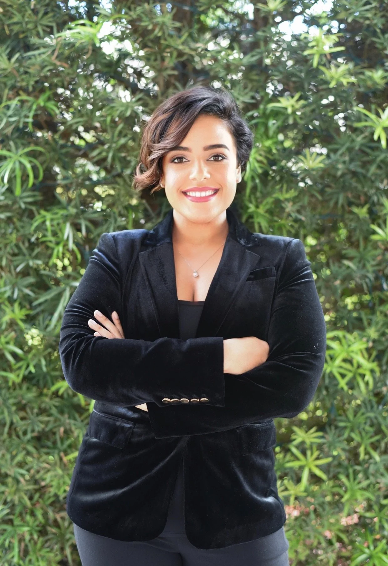 A woman with short dark hair, wearing a black blazer and black top, stands with arms crossed in front of green foliage, smiling.