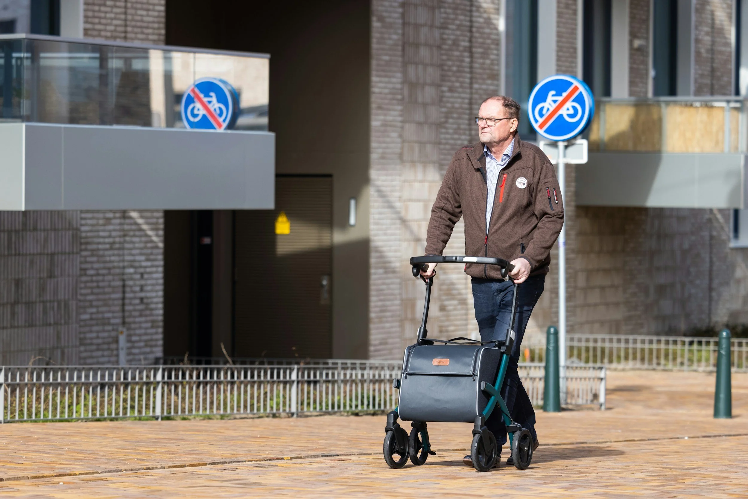 A man with glasses walking outside during the day, pushing a walker with a foldable seat, on a brick paved walkway near a building with brick walls and large windows. There are blue signs indicating no bicycles.