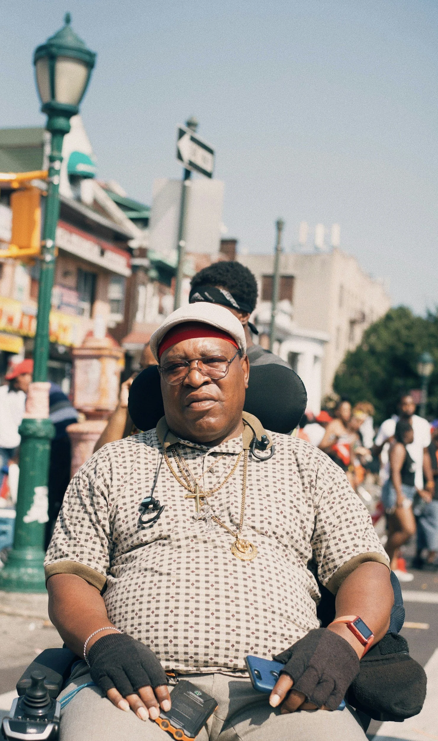 Man with glasses and jewelry, sitting in a wheelchair outdoors, wearing a patterned shirt, black gloves, and a headband.