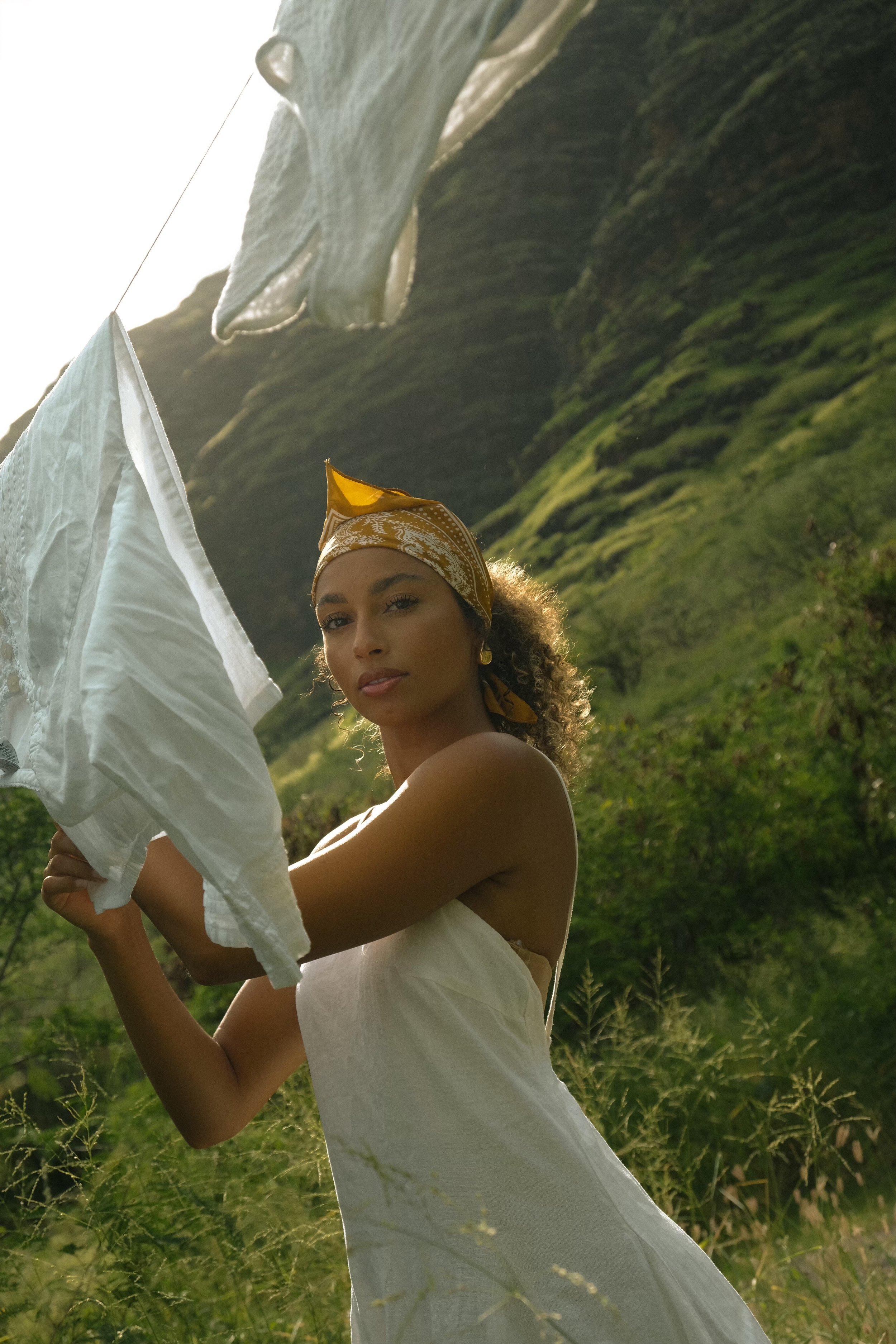 A woman with curly hair and a yellow headscarf standing outdoors on a lush green hillside, holding white fabric or paper, with mountains and bright sky in the background.