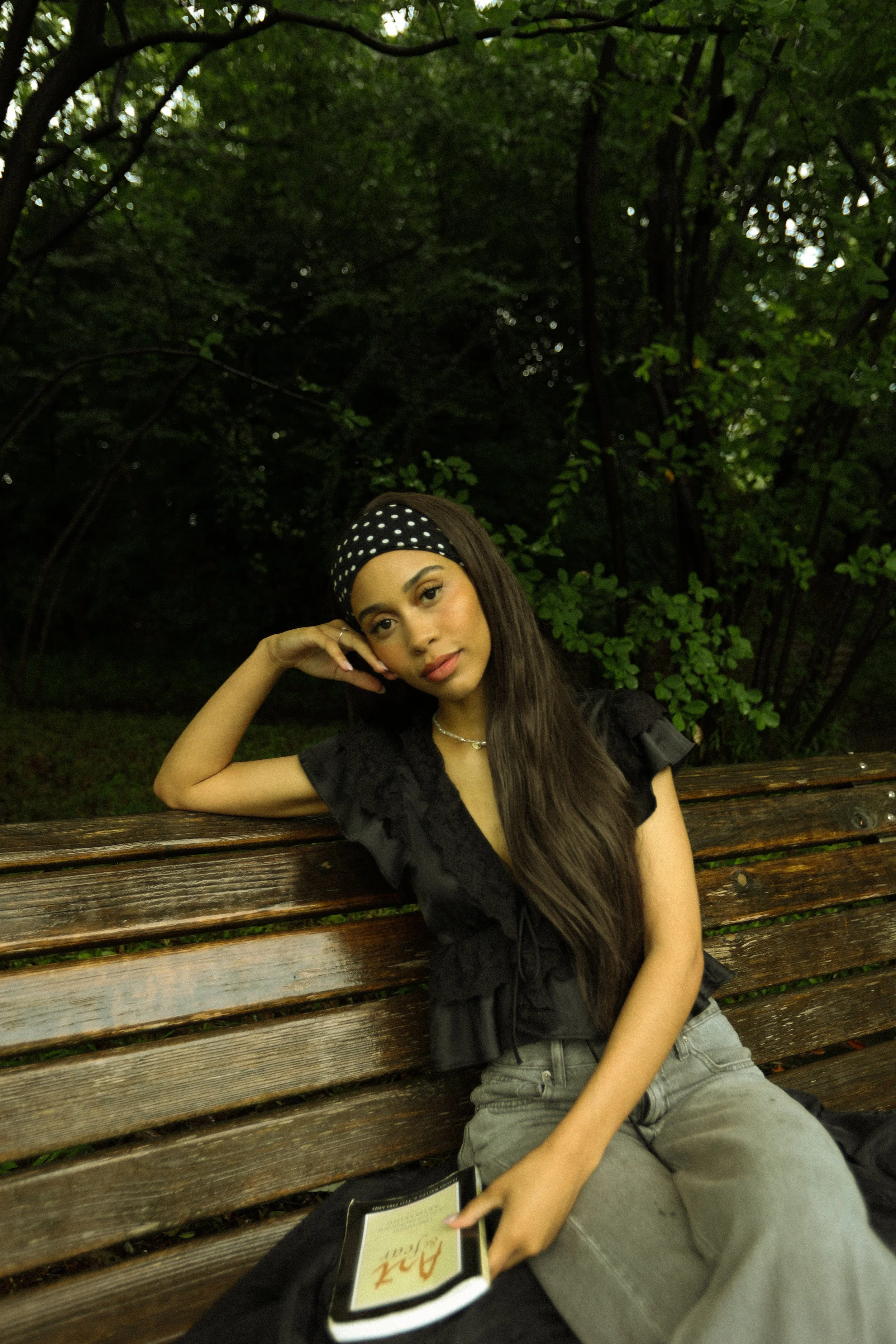 A young woman with long hair, leaning on a park bench, wearing a black headband with white polka dots, a black top, and gray jeans, sitting outdoors in a wooded area with green trees in the background.