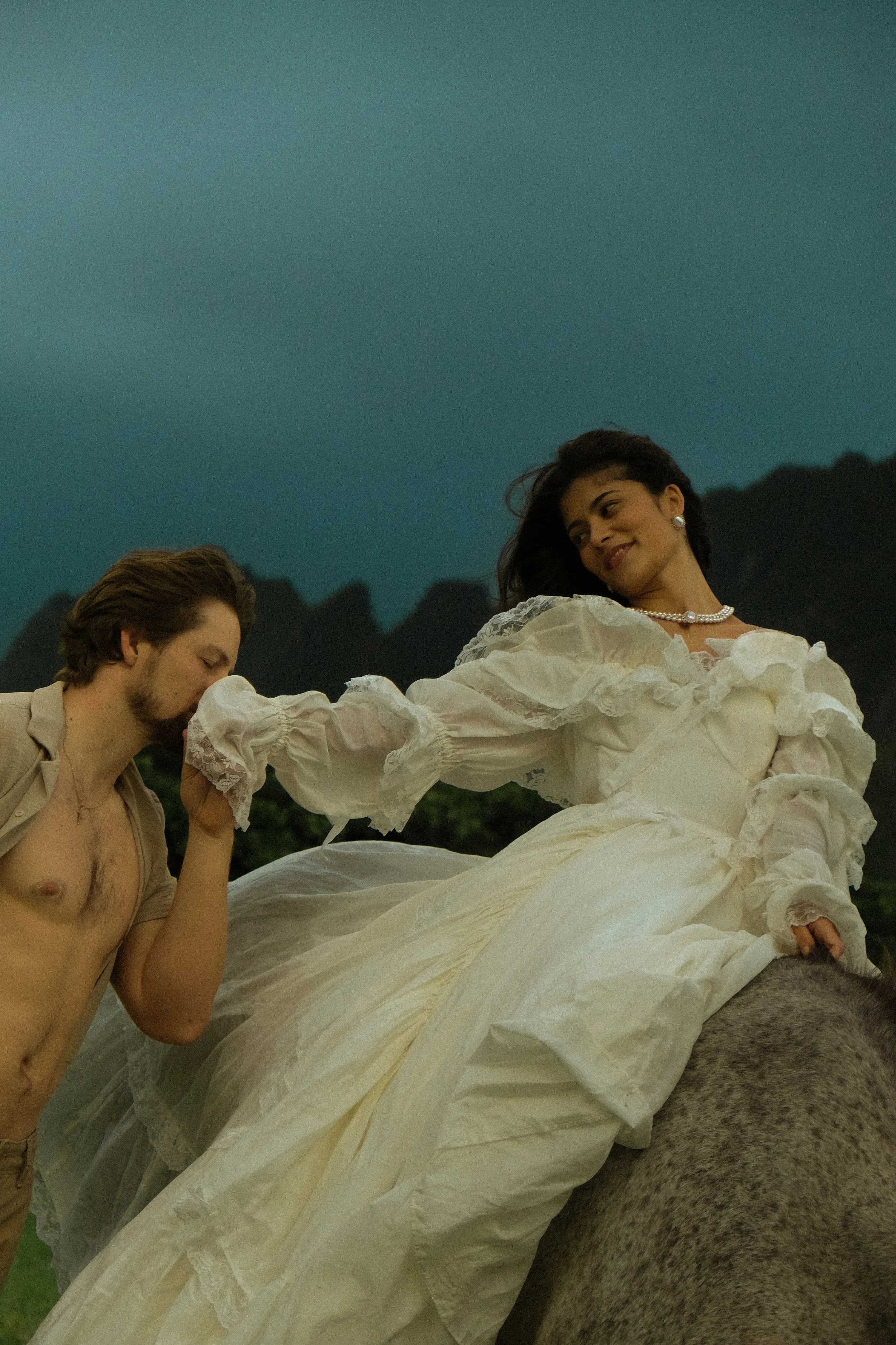 A woman in a white vintage dress with puffed sleeves and pearl necklace sitting on a horse, escorted by a shirtless man with tousled hair and beard, against a mountain landscape at dusk.