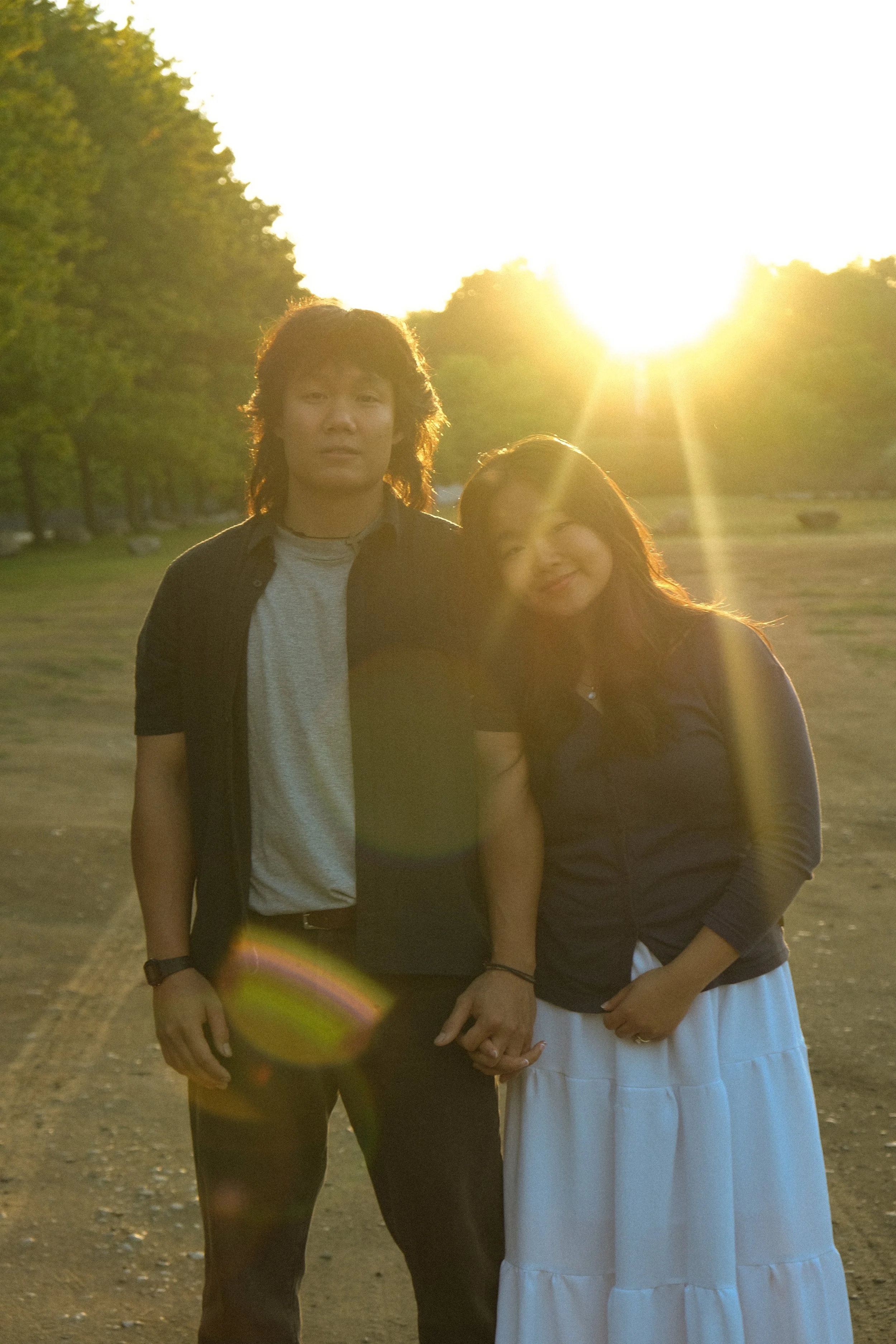 A man and woman standing hand in hand outdoors during sunset, with trees and a field in the background.