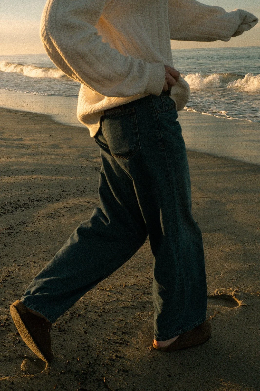 Person standing on a beach at sunset, wearing a chunky sweater, loose jeans, and brown shoes, with ocean waves in the background.