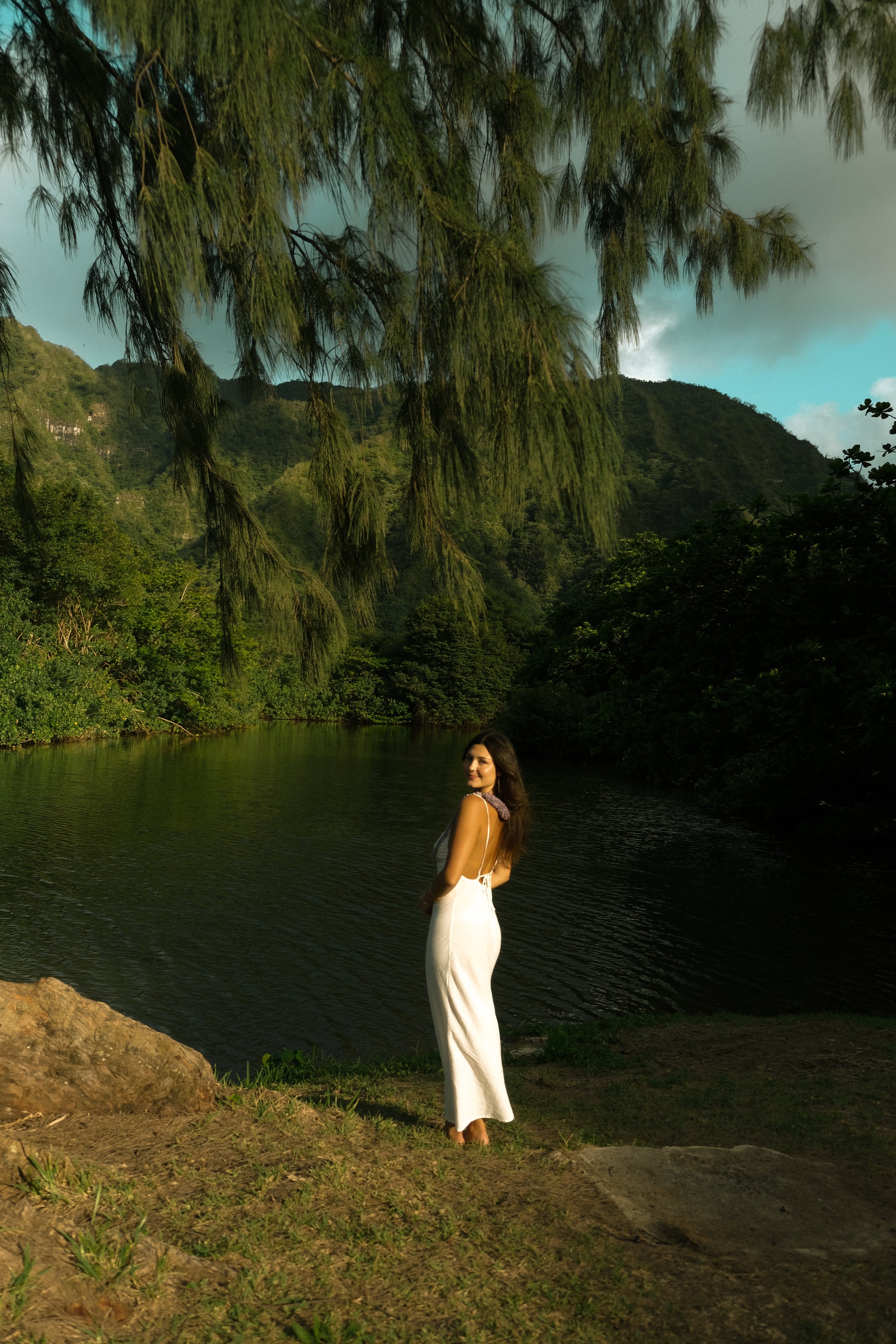 A woman in a white dress standing near a river with trees and mountains in the background.