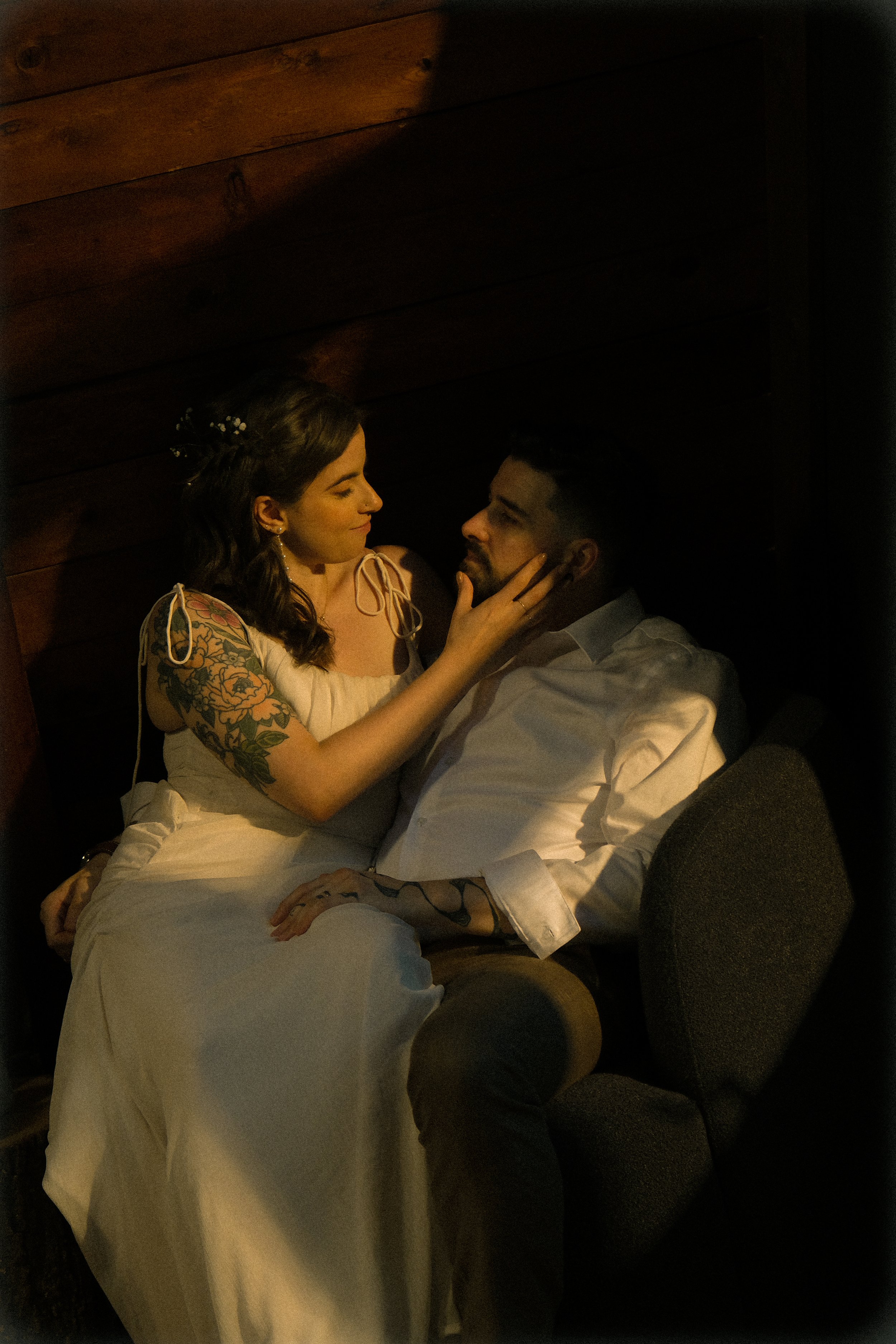 A woman with tattoos, wearing a white dress, gently touching a man's face. The man is sitting, dressed in a white shirt, with dark hair and a beard, looking at her. They are in a dimly lit room with wooden walls.
