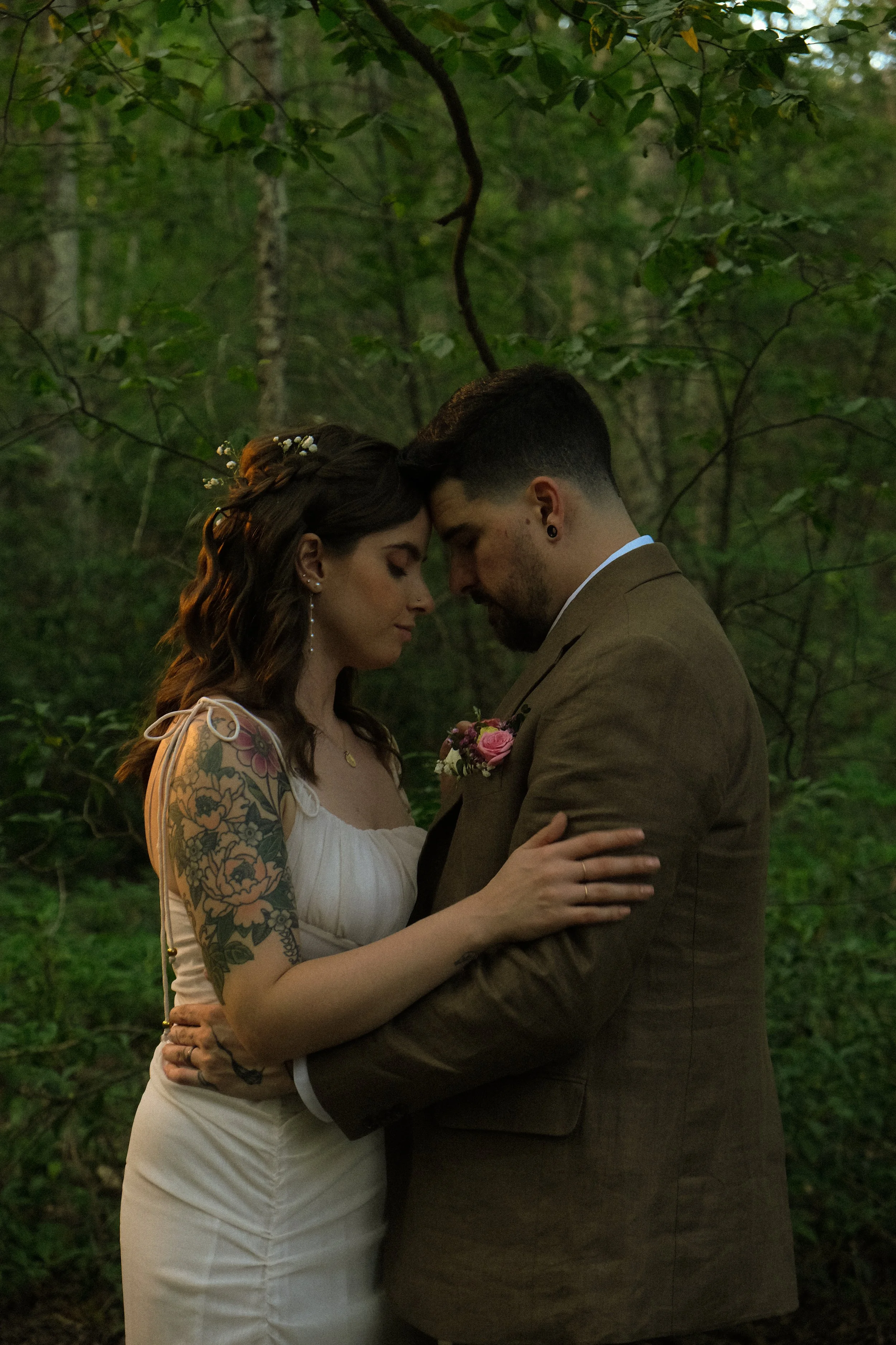 A couple in wedding attire embracing in a forest, with their foreheads touching and eyes closed.