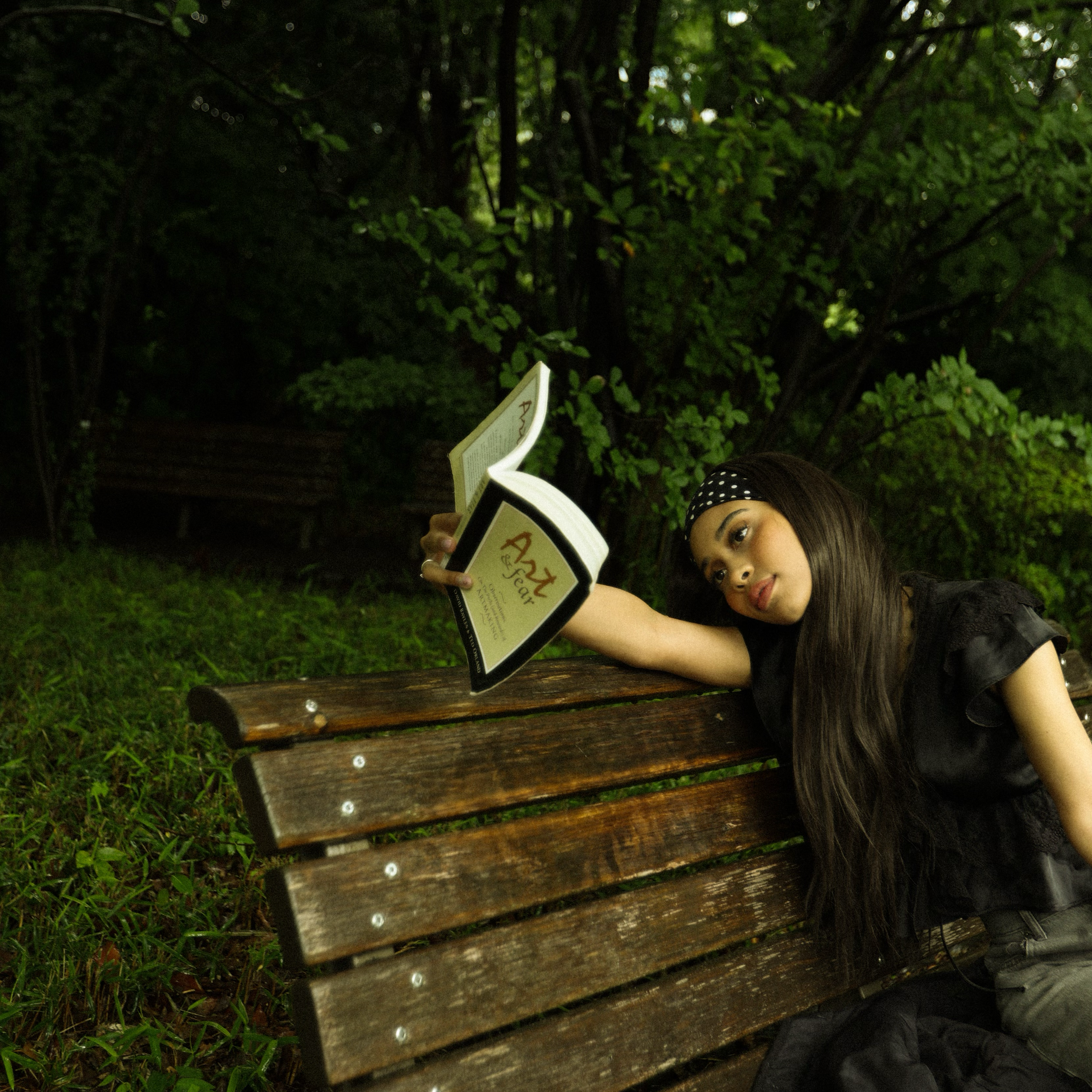 A woman with dark hair and a polka dot headband stares at the camera while sitting on a wooden park bench with lush green trees in the background, holding a book.