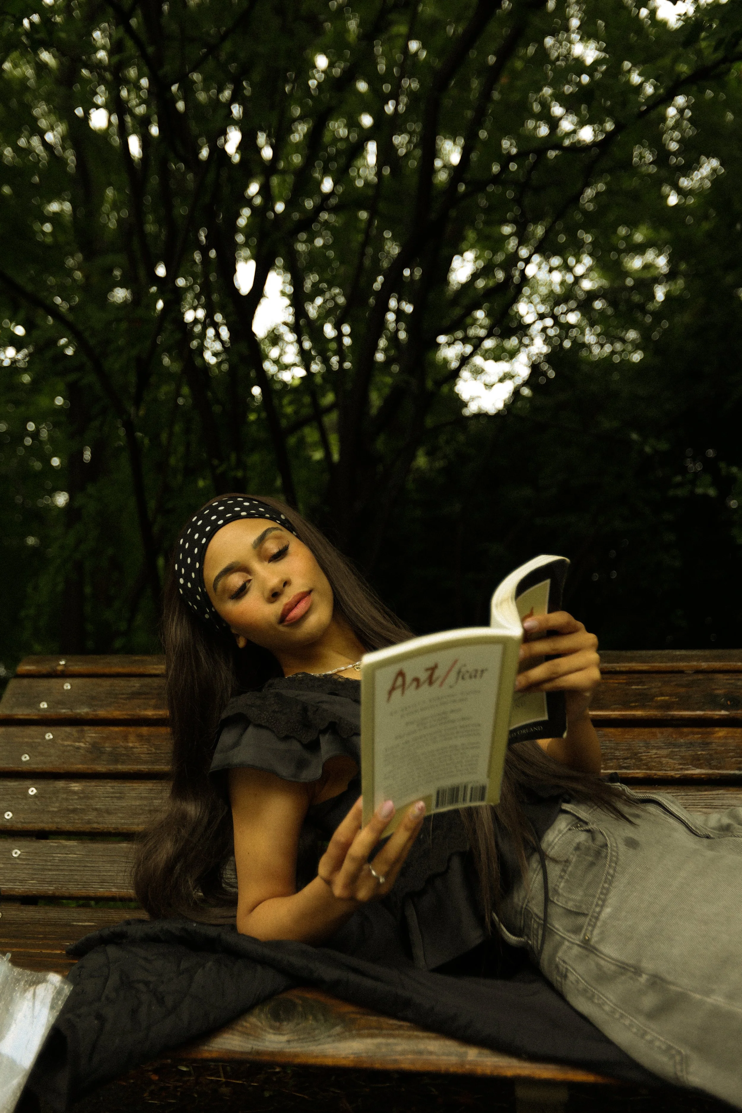A young woman with long dark hair, wearing a black headband with white polka dots, is lying on a wooden park bench. She is reading a book titled 'Art/Sciar'. She is dressed in a black ruffled top and beige pants, with a black coat spread beside her. 